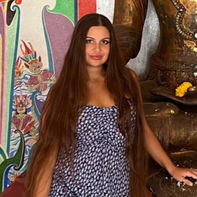 A woman with long brown hair, wearing a patterned dress, standing in front of a colorful mural with a Thai theme and a large Buddha statue.