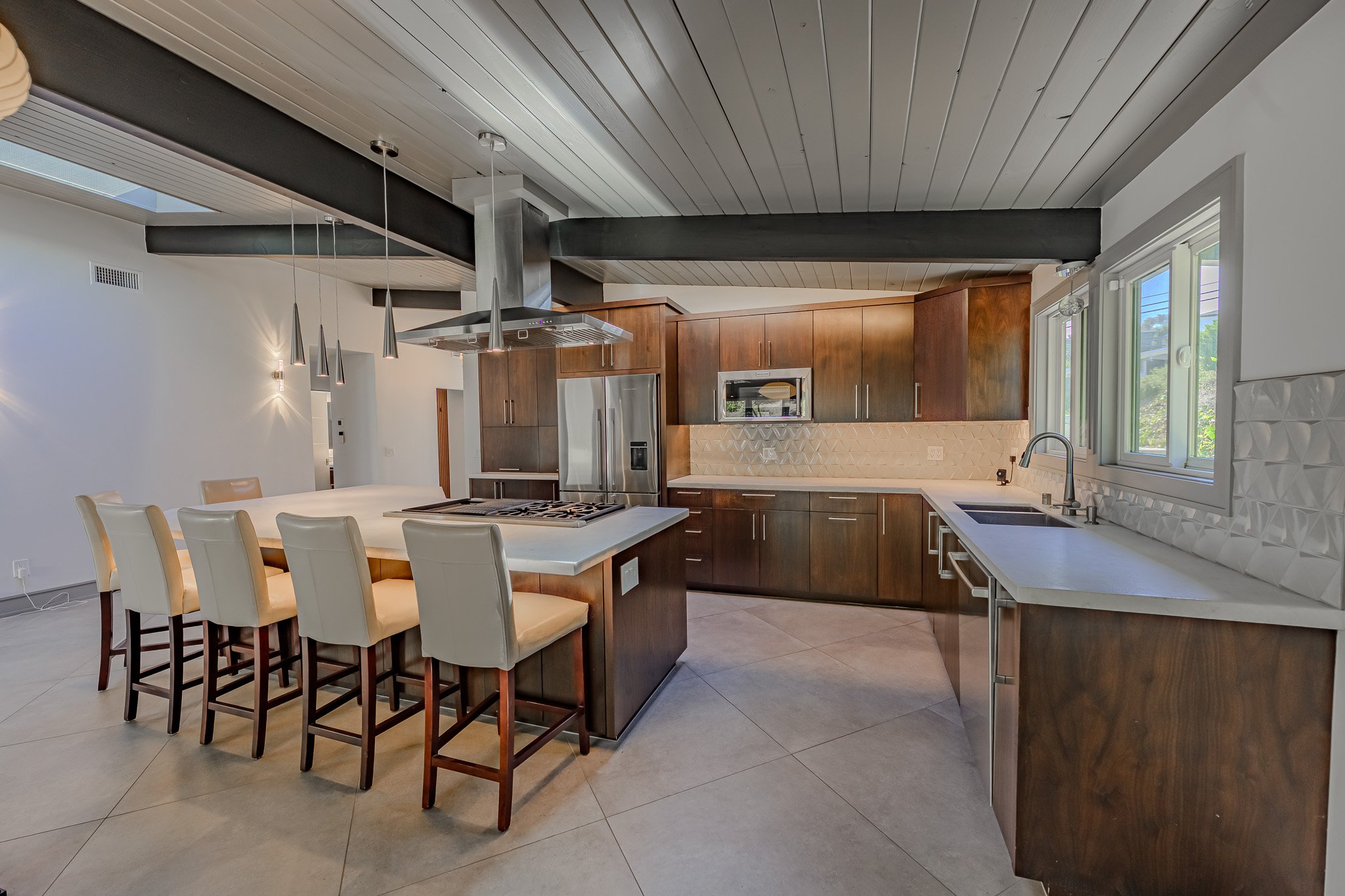 Modern kitchen with white and wood cabinetry, stainless steel appliances, a center island with a stovetop, and beige chairs around a kitchen counter. Large windows provide natural light.