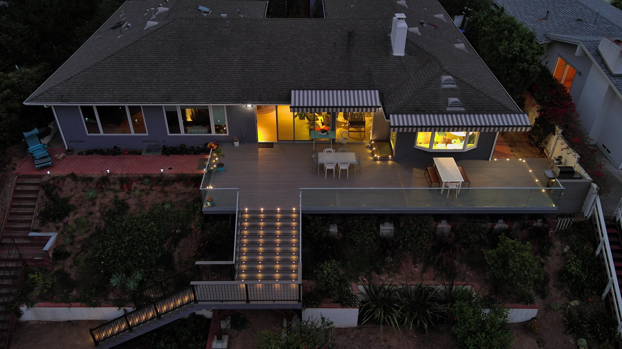 Aerial view of a house with a large wooden deck, illuminated outdoor lighting, patio furniture, and a staircase leading down to a garden area, during dusk or early evening.
