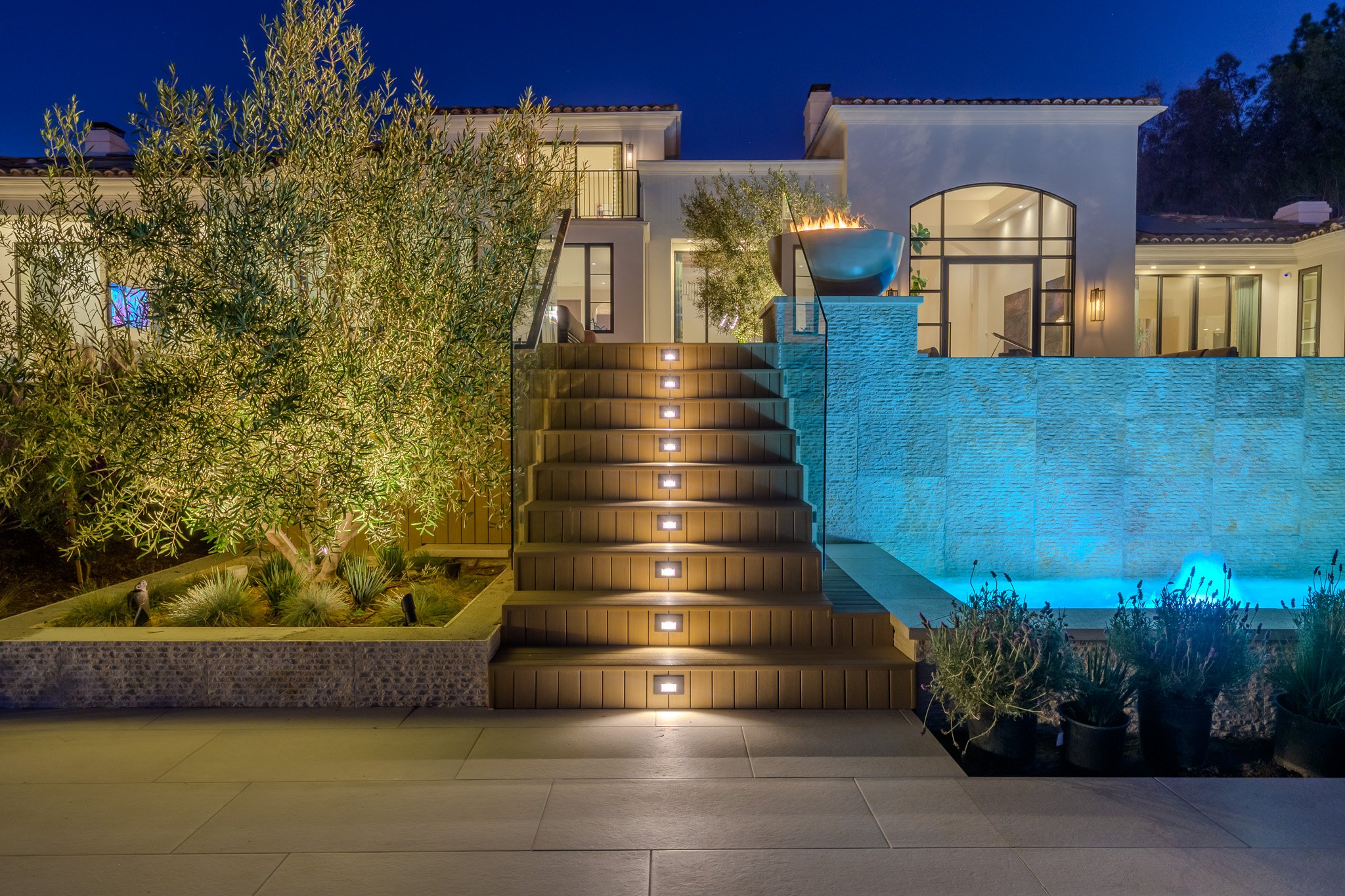 Night view of a modern backyard with a swimming pool, illuminated stairs, outdoor fire bowl, landscaped plants, and a white house with large windows.