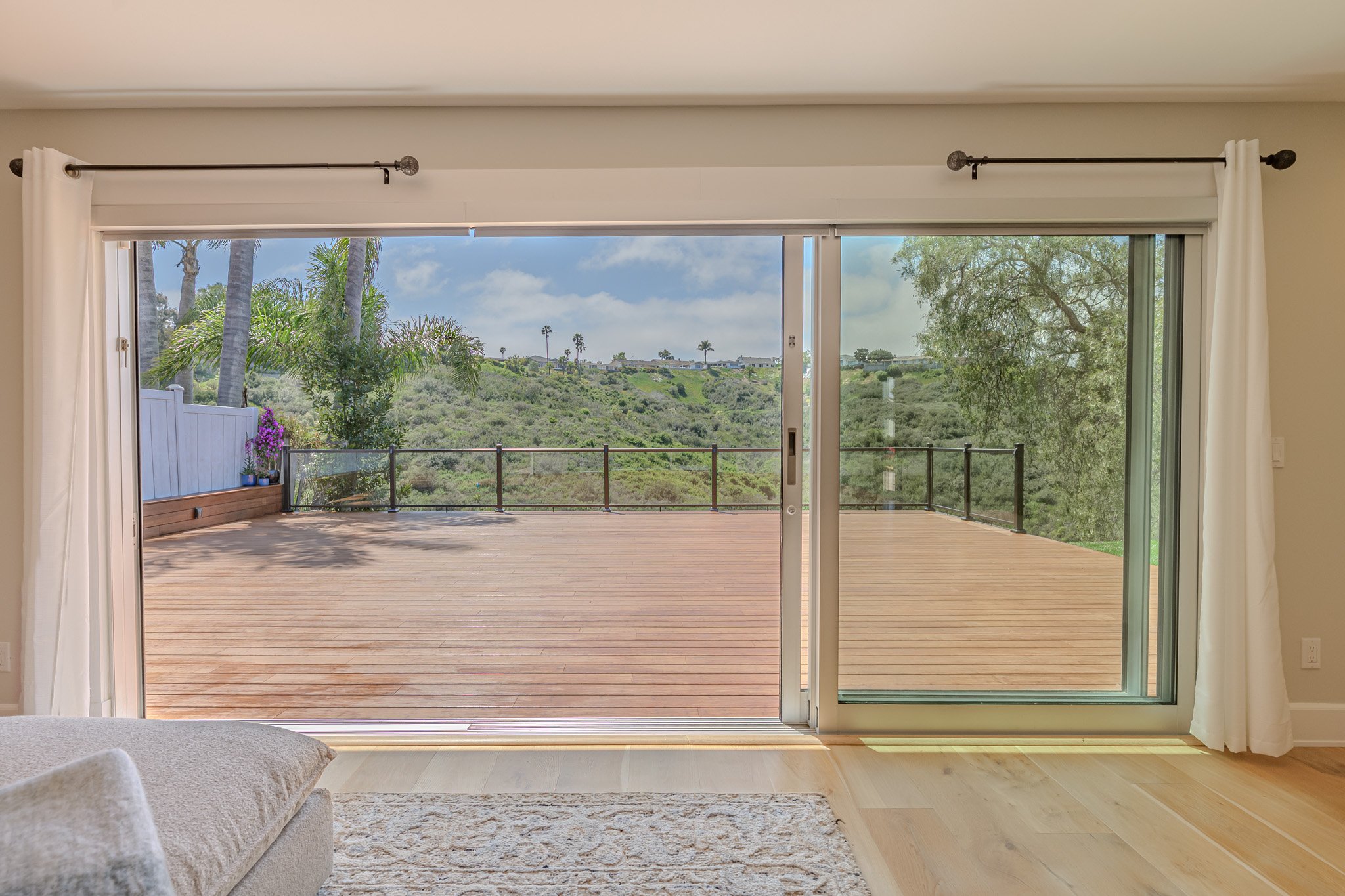 Living room with sliding glass doors opening to a wooden deck and a lush hilly landscape with palm trees outside.