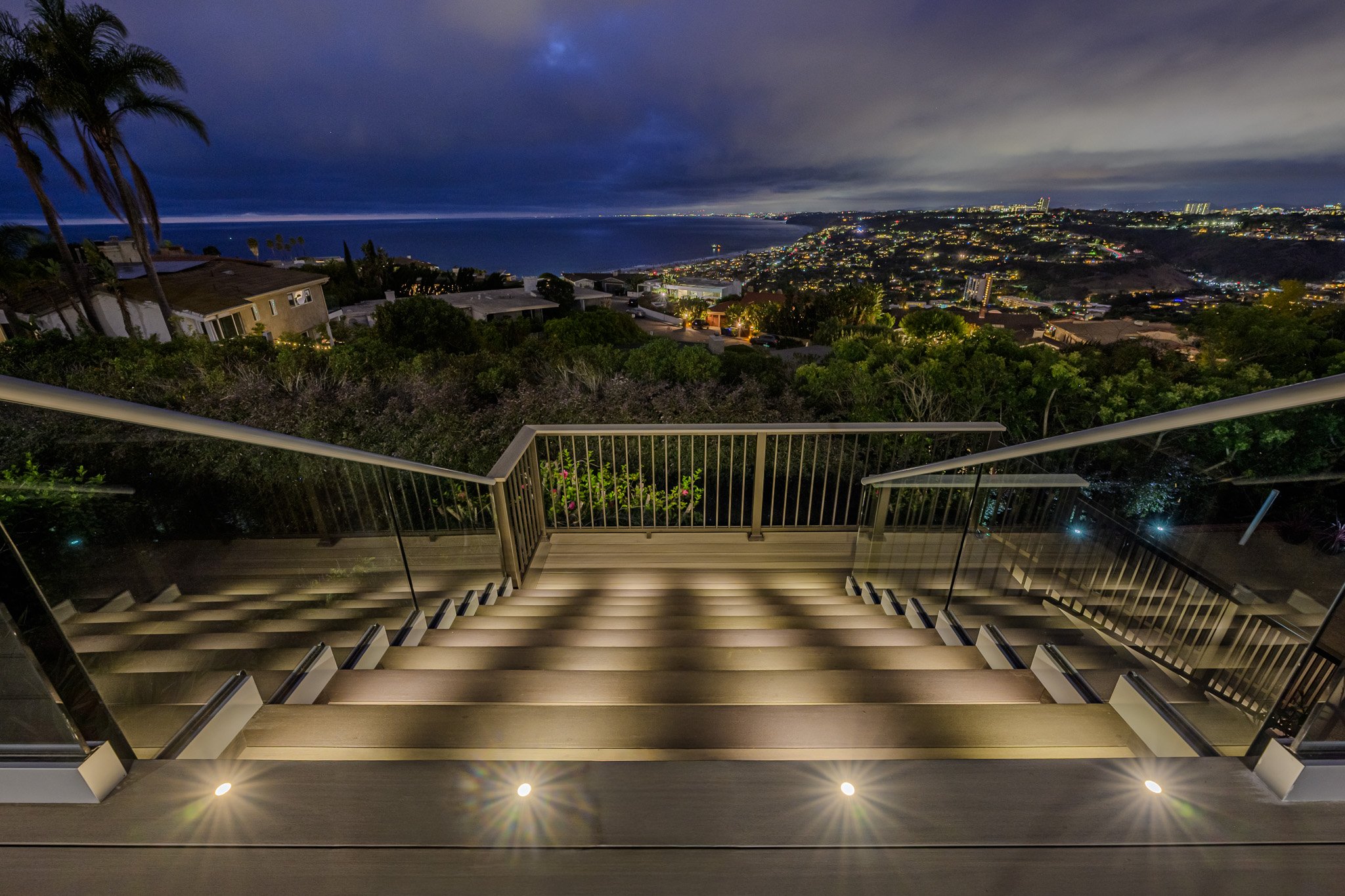 Night view from a balcony with stairs, overlooking a city and ocean under a cloudy sky, illuminated city lights and palm trees visible.