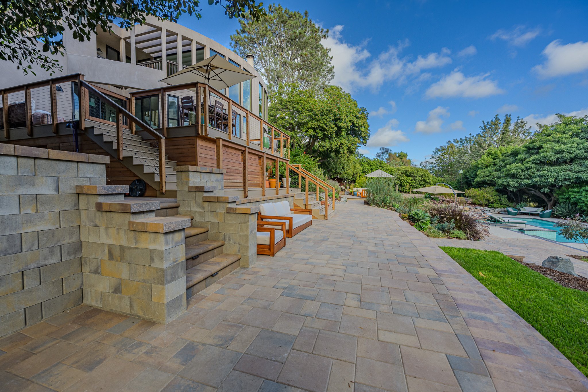 A backyard with a patio, pool, and lush green trees under a blue sky with some clouds. There is outdoor seating, a wooden deck with stairs, and a house with large windows.