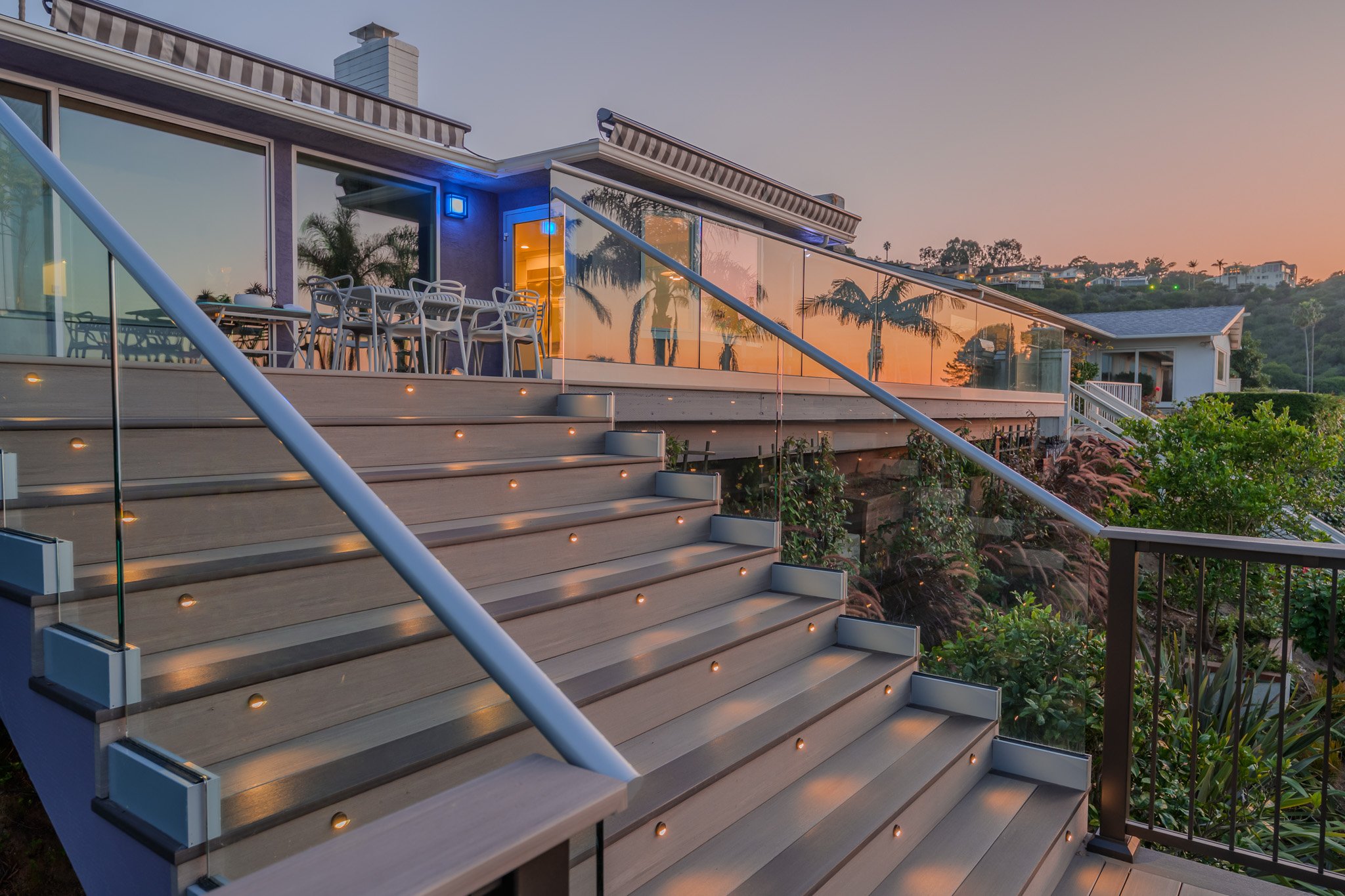 View of a modern house with a glass railing balcony, outdoor dining table with chairs, and surrounding greenery during sunset.