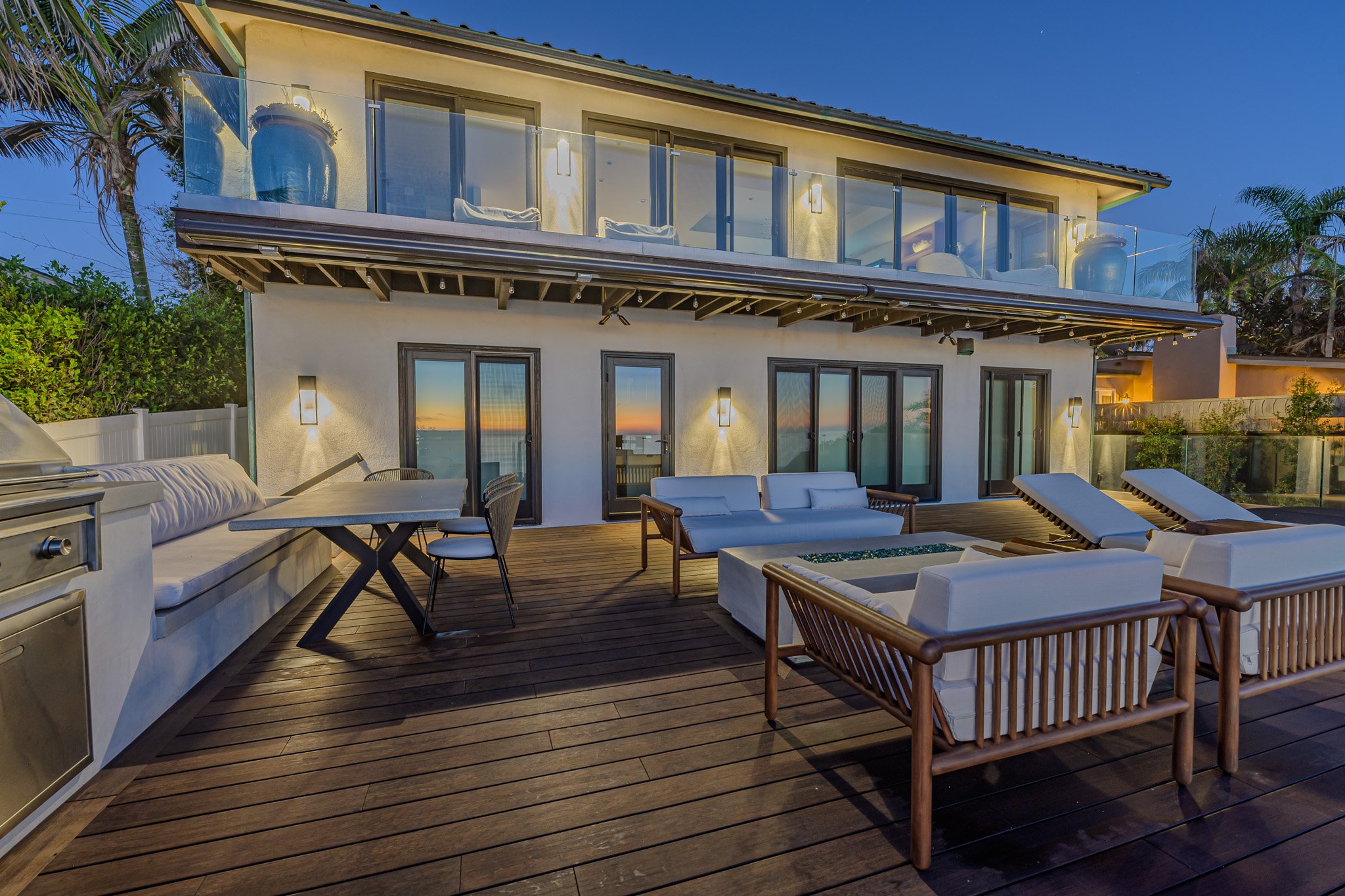 A modern two-story house with a spacious outdoor deck, furnished with white sofas, chairs, a dining table, and a barbecue grill, overlooking the ocean at sunset. The house has large sliding glass doors, a glass balcony, and is surrounded by palm trees.