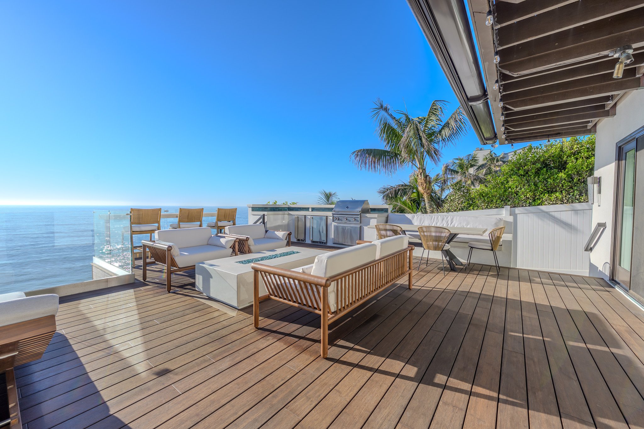 Modern outdoor patio with wooden flooring, white and wooden outdoor furniture, palm trees, and ocean view, under a clear blue sky.
