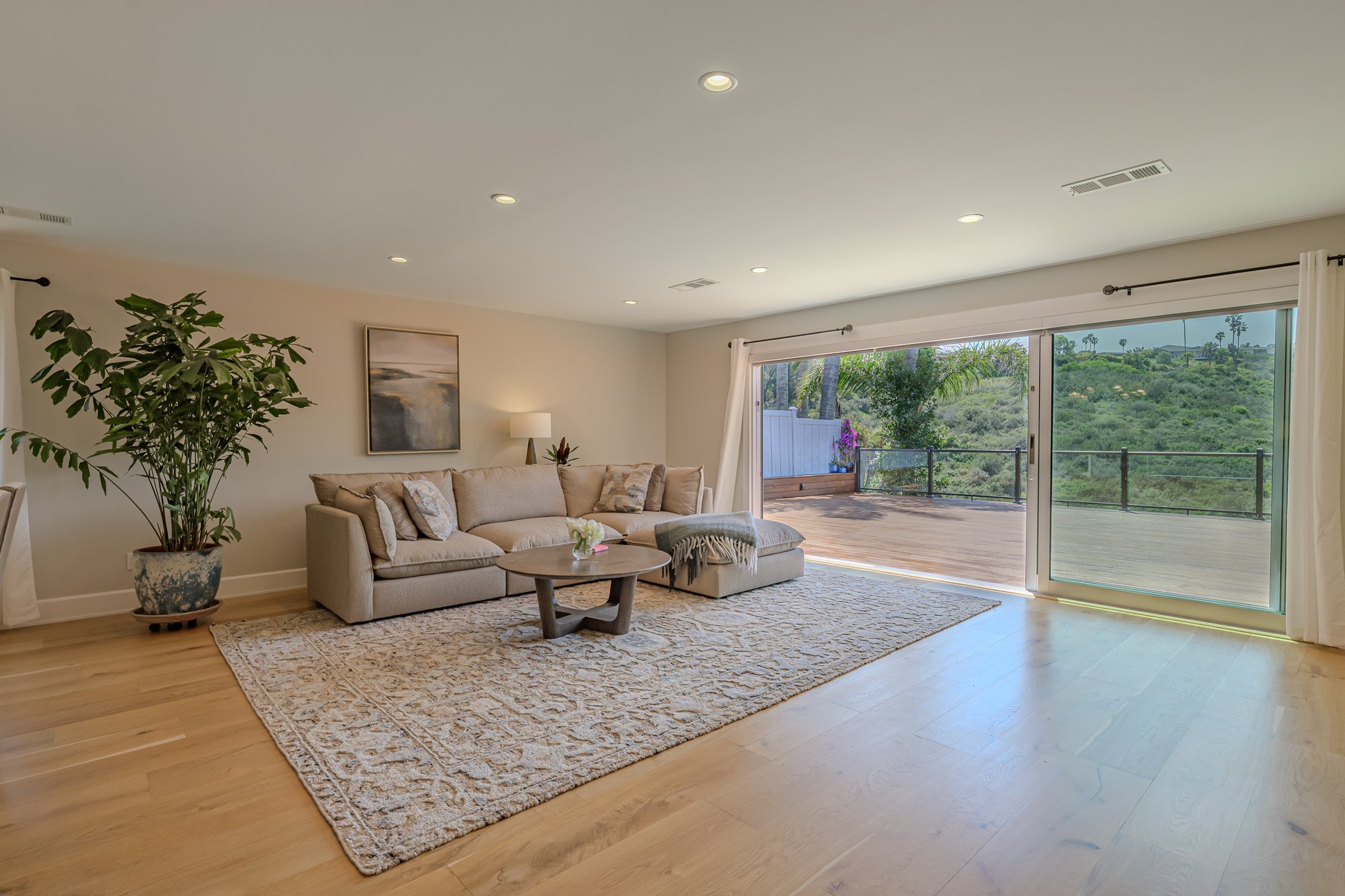 Living room with beige sofa, large potted plant, coffee table, artwork on wall, and sliding glass door leading to outdoor patio with greenery and trees.