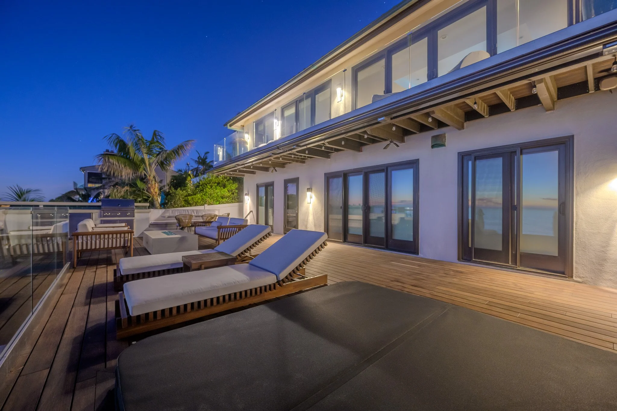 Nighttime view of a modern balcony with lounge chairs, a dining table, and a railing, overlooking the water. The building has large glass windows and doors, with exterior lighting and palm trees in the background.