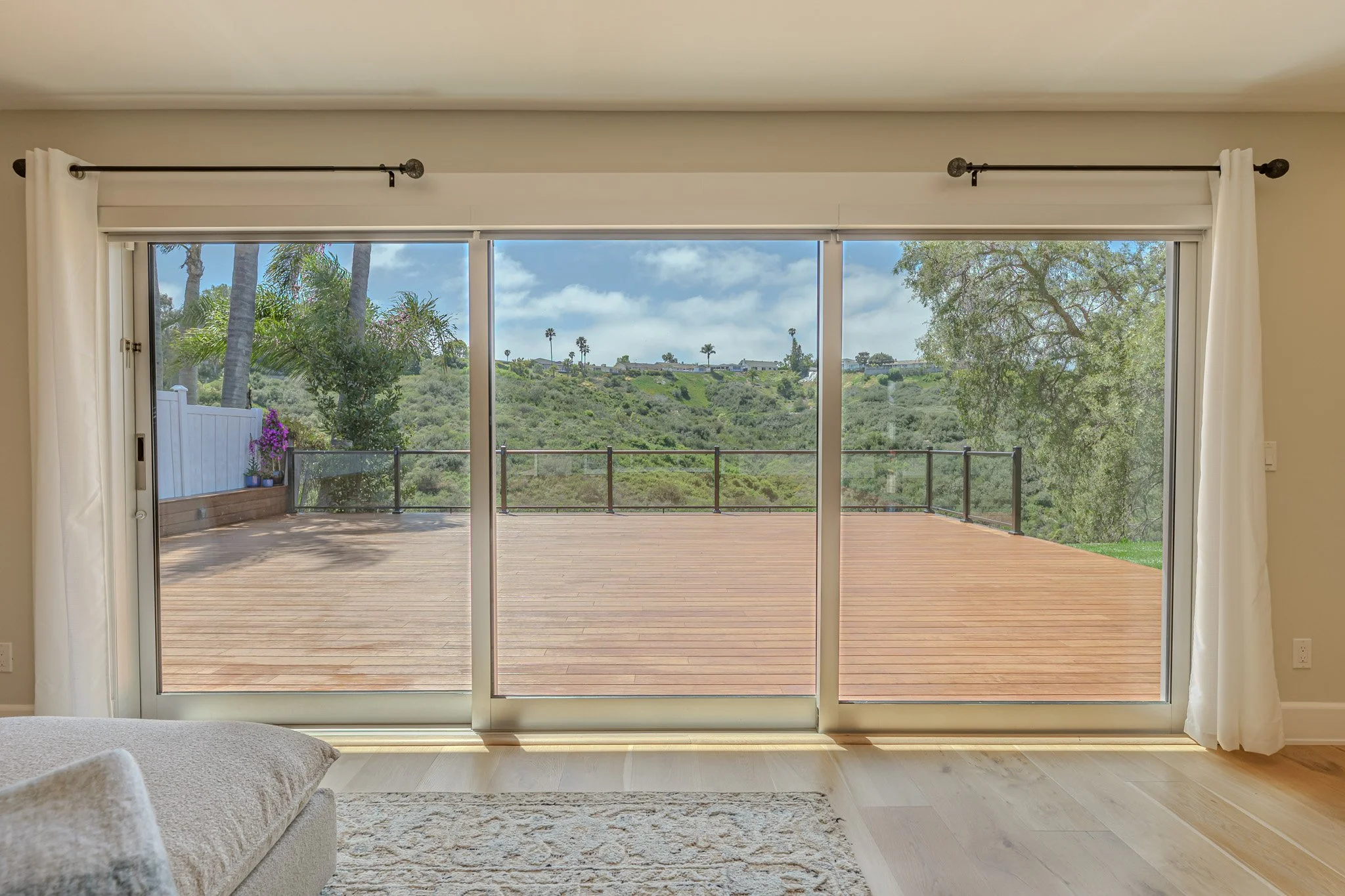 View through sliding glass doors showing a wooden deck, greenery, and hilly landscape with trees and houses in the distance under partly cloudy sky.