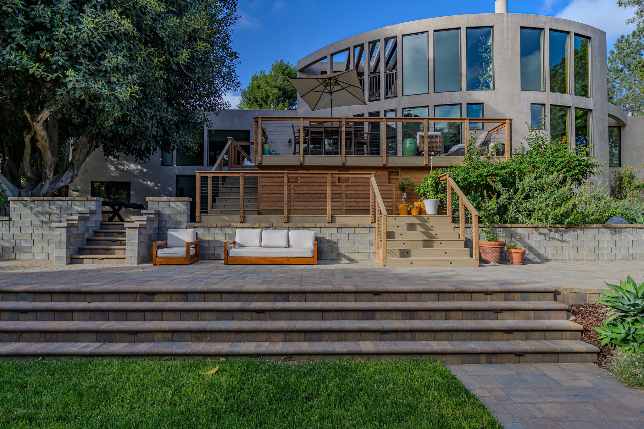 Modern multi-story house with large glass windows and a wooden deck, outdoor furniture including a sofa and chairs, and a staircase leading up to the deck. Green bushes and trees are surrounding the house, with a bright blue sky overhead.