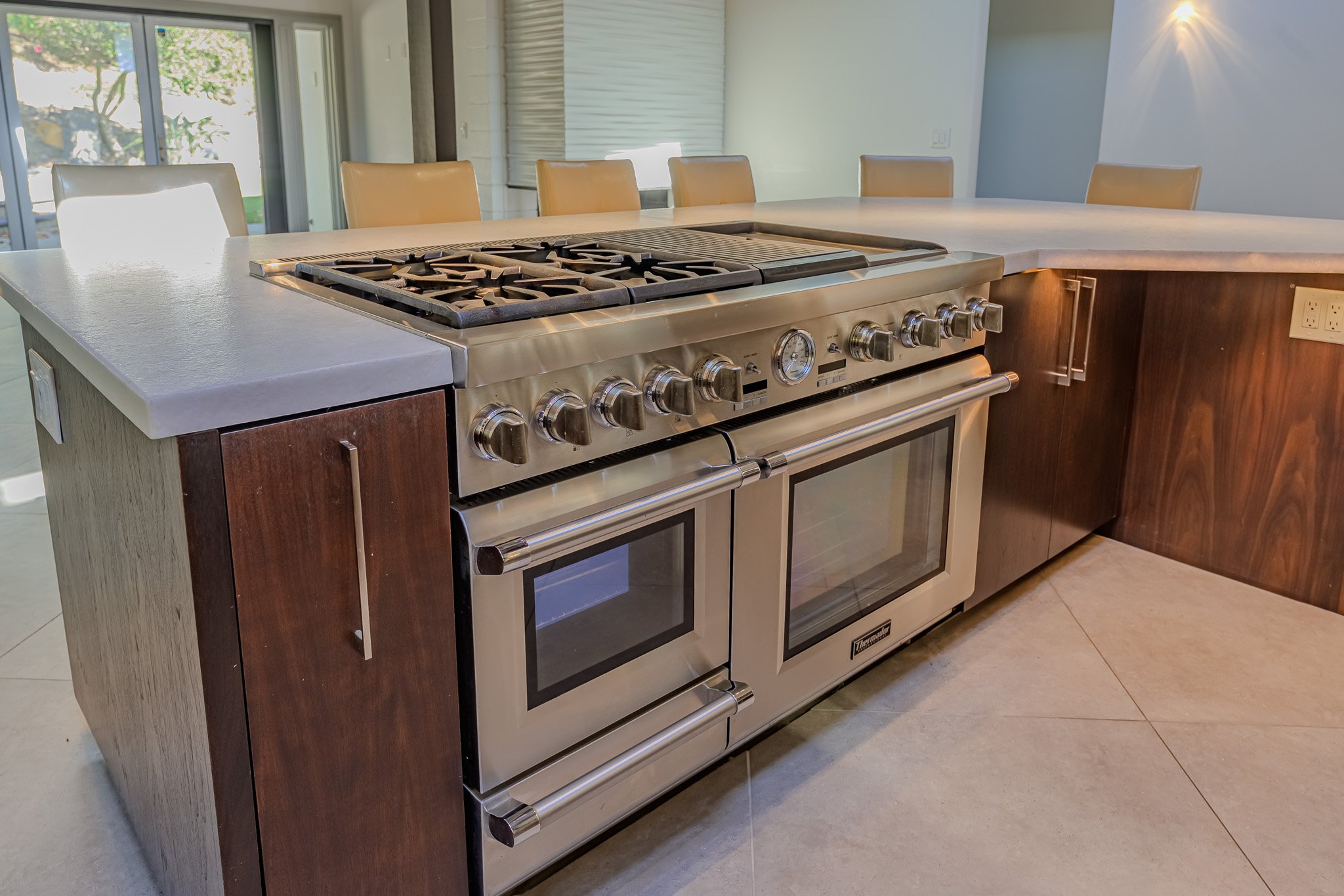 Stainless steel gas range stove with six burners and a built-in oven, set in a kitchen with tiled flooring and surrounded by a white counter and wooden cabinetry.