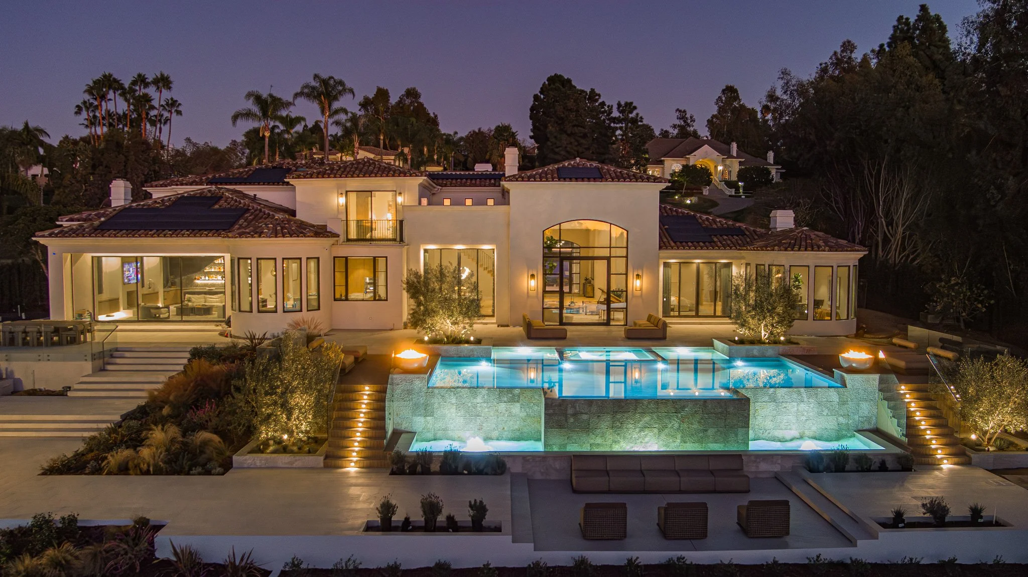 Luxury house with a swimming pool and outdoor seating area illuminated at night, surrounded by trees and landscaping.