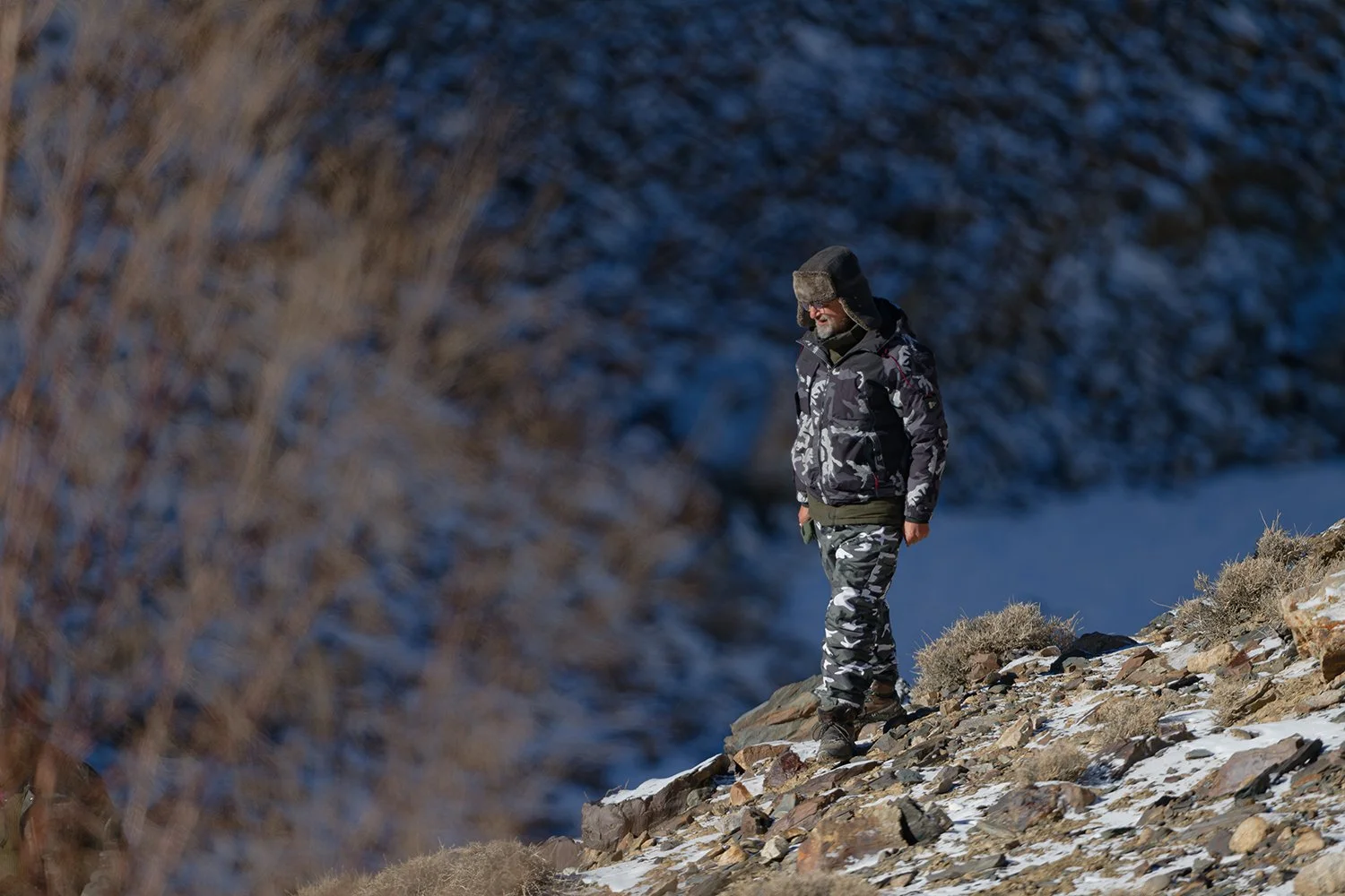 A man wearing camouflage clothing and a fur-lined trapper hat walking on rocky terrain in a snowy, mountainous landscape.