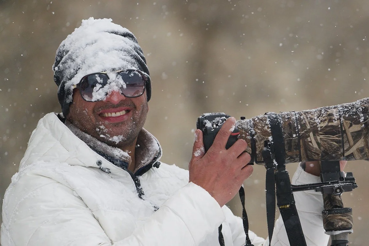 A man wearing sunglasses, a black beanie, and a white winter jacket is smiling while holding a camera with a camouflage lens cover. Snow is falling around him.