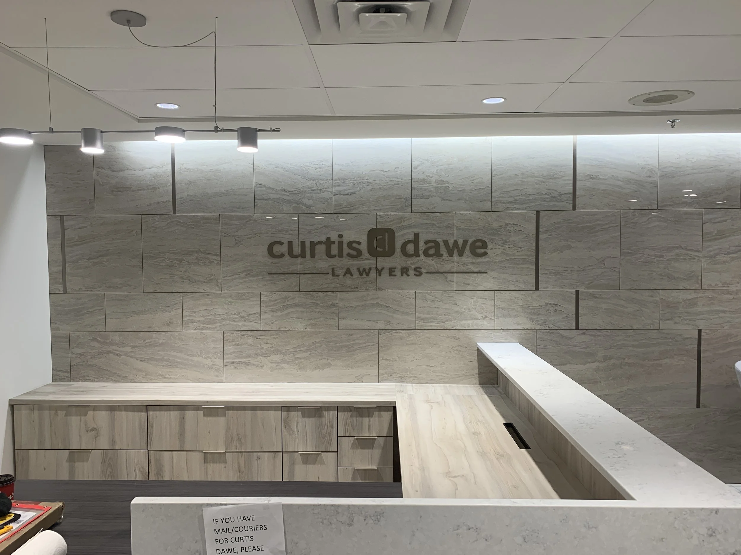 Reception area of Curtis Dawe Lawers office with a marble wall and logo, a light-colored wooden counter, and overhead lighting.