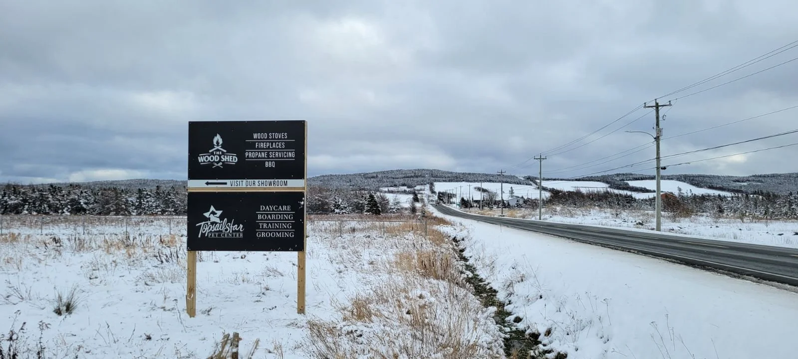 Snow-covered roadside with a black signboard advertising wood stoves, fireplaces, propane servicing, BBQ, and a pet center, near a road with utility poles, in a rural area with distant snow-covered hills.