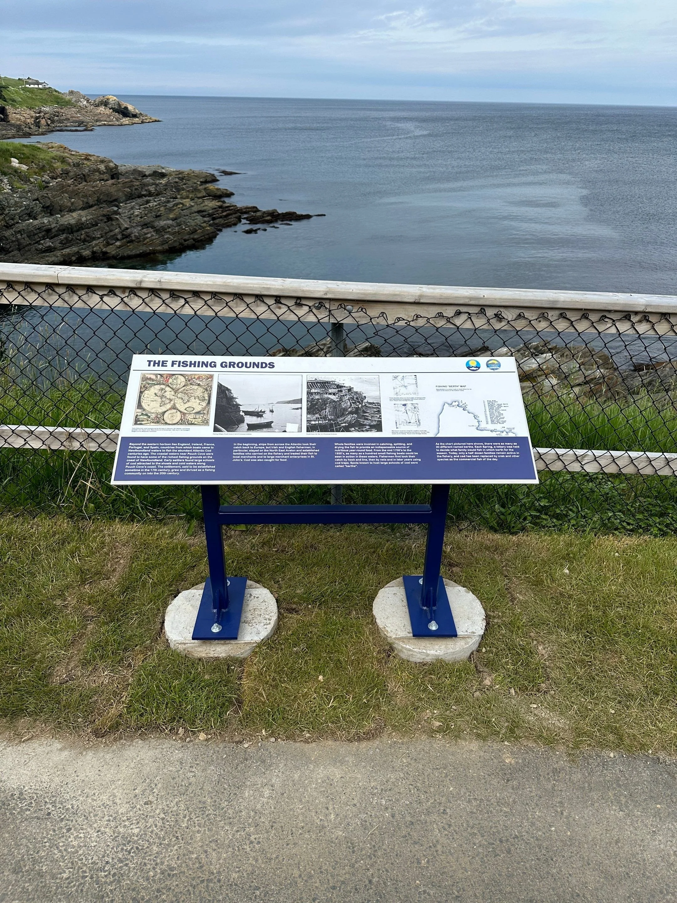Information board titled 'The Fishing Grounds' in front of a coastal view with rocks and water, enclosed by a fence.