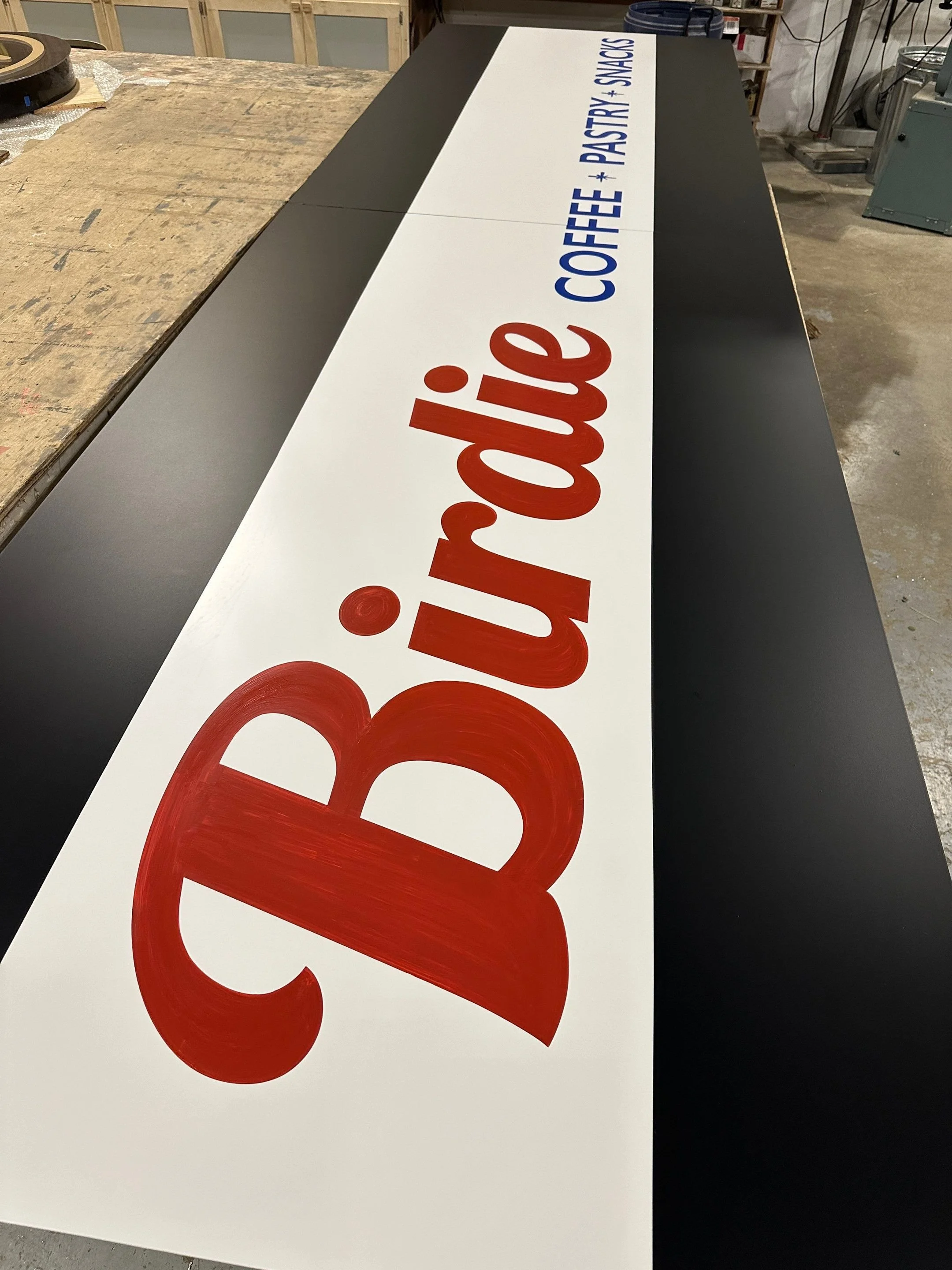 A black and white sign with red and blue lettering reading 'Bev's Deli'. The sign is positioned on a worktable in what appears to be a workshop or storage area.