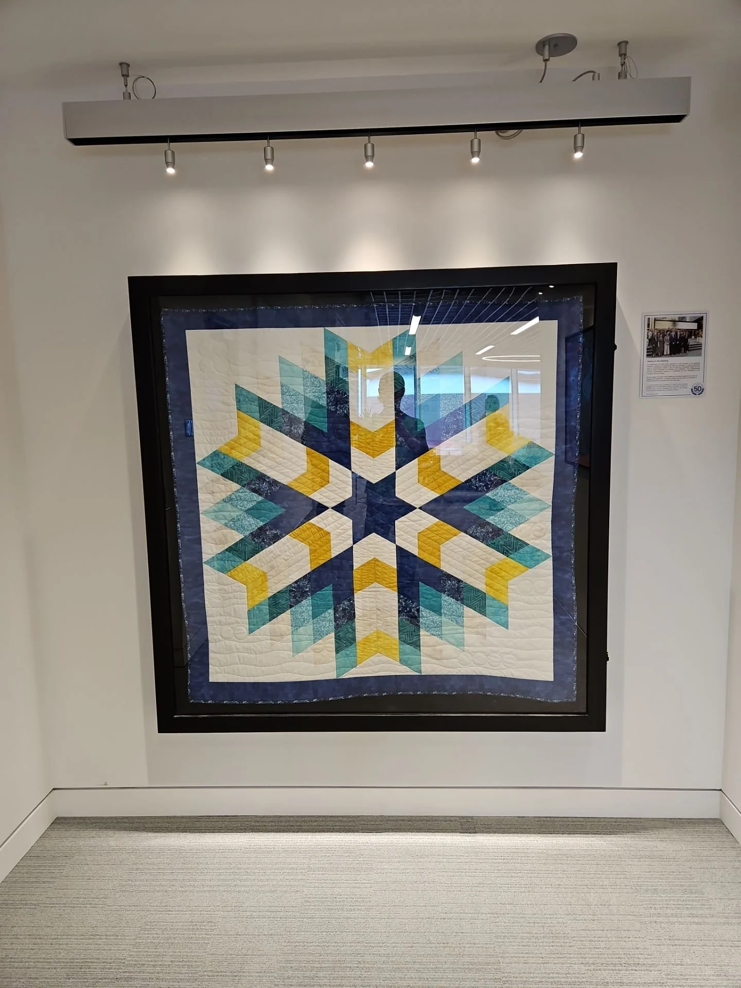 A colorful quilt with a snowflake pattern, framed and displayed on a wall, with a reflection of the room in the glass.