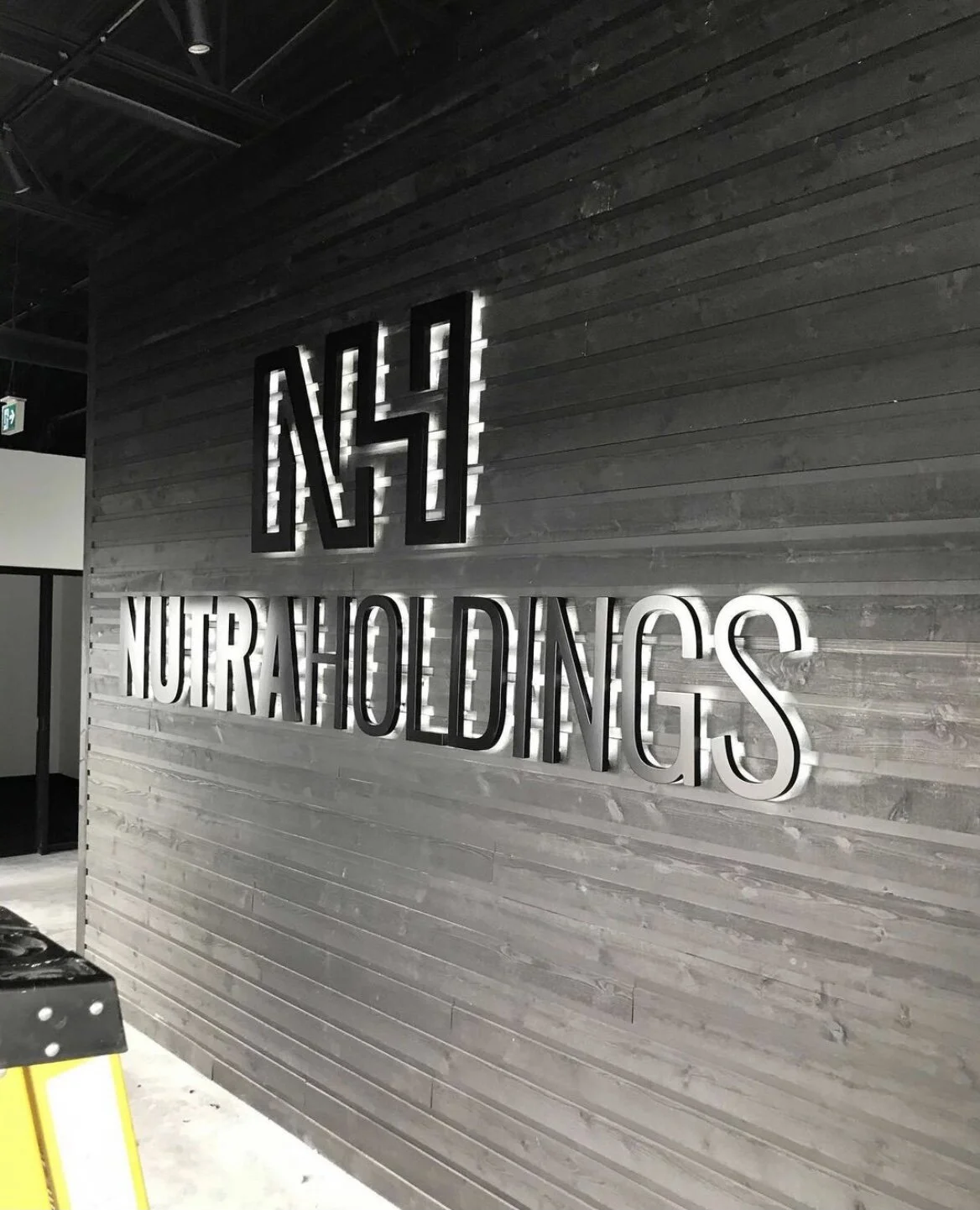 A gray wooden wall with illuminated black and white signage reading 'NH NUTRA HOLDINGS'.