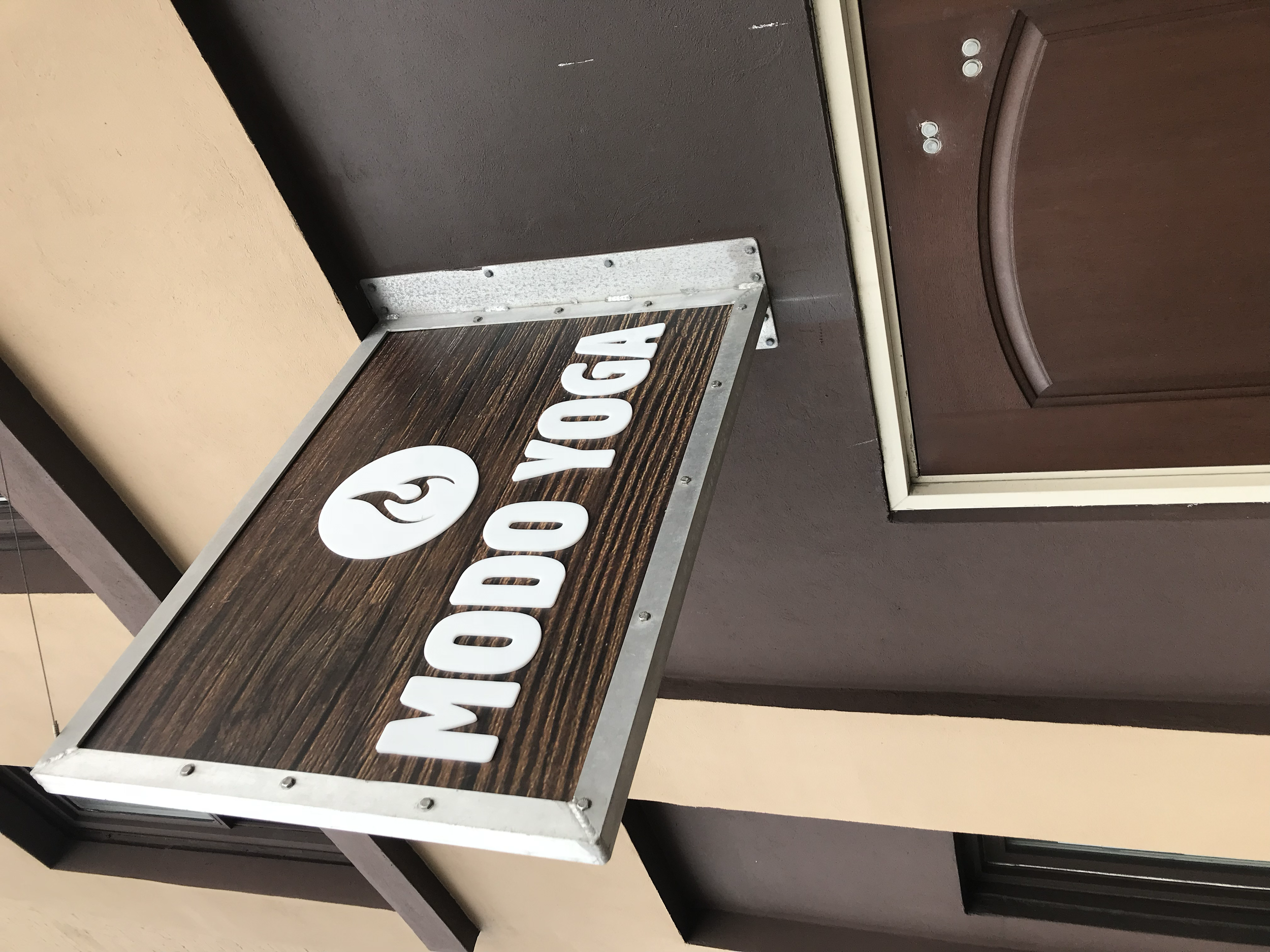 Wooden sign with white text saying 'MODO YOGA' and a circular logo, attached to a building exterior wall near a brown door.