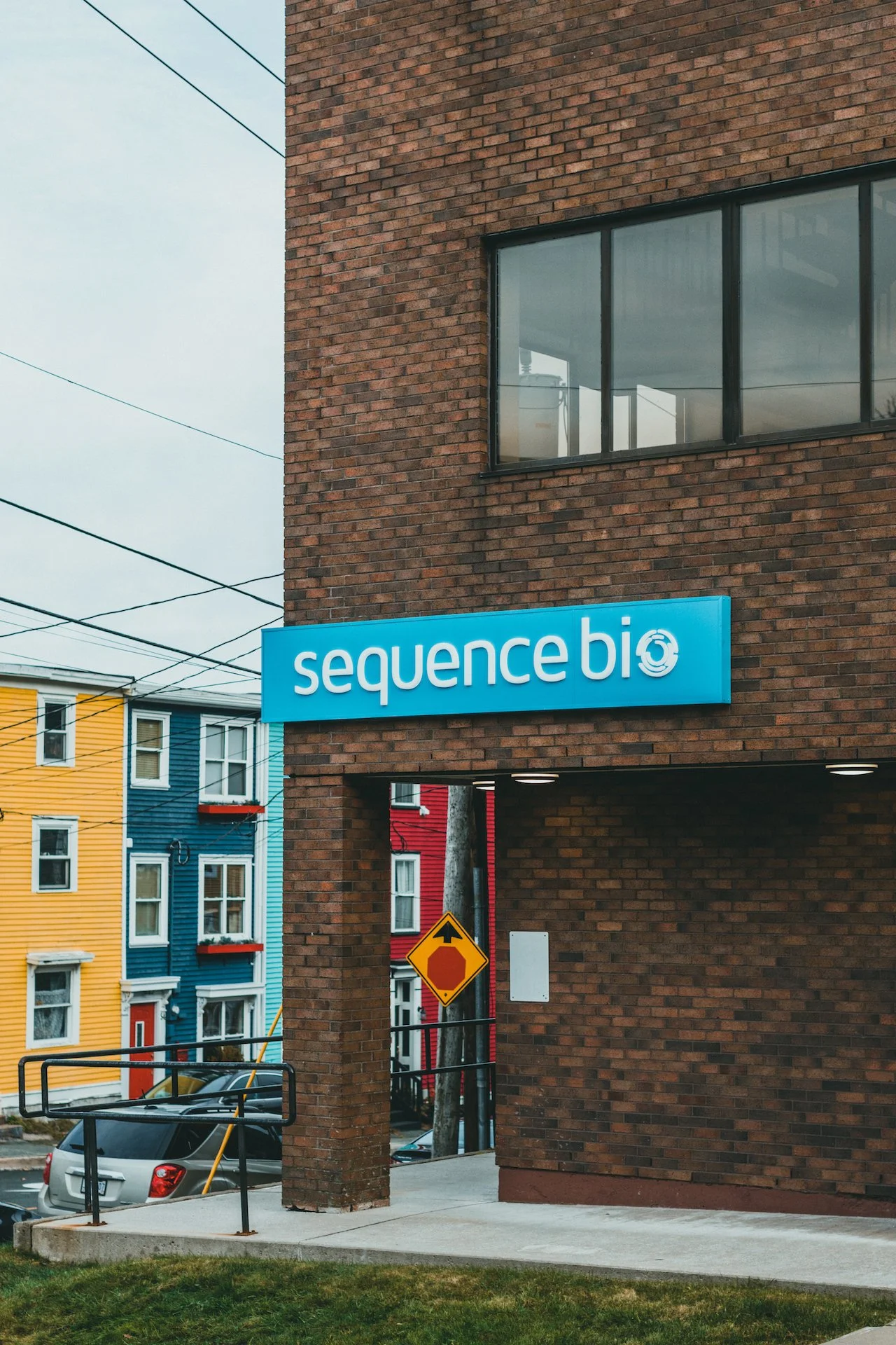 A blue sign with white lettering reading 'sequence bio' on a brick building. There is a yellow and red pedestals sign, a gray car parked nearby, and colorful houses in the background.