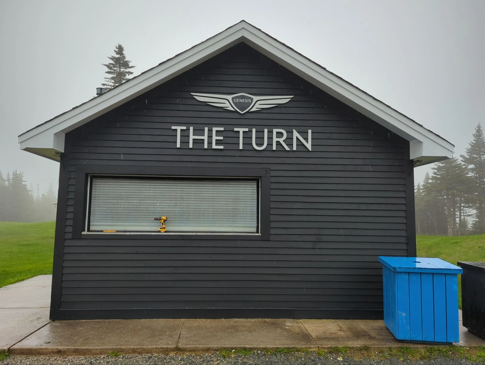 A black wooden building with the sign 'The Turn' and a logo 'Genesis' above it, a yellow power drill on the window ledge, and two trash bins, one blue and one black, outside on a paved sidewalk with a grassy field and trees in the background on a fog