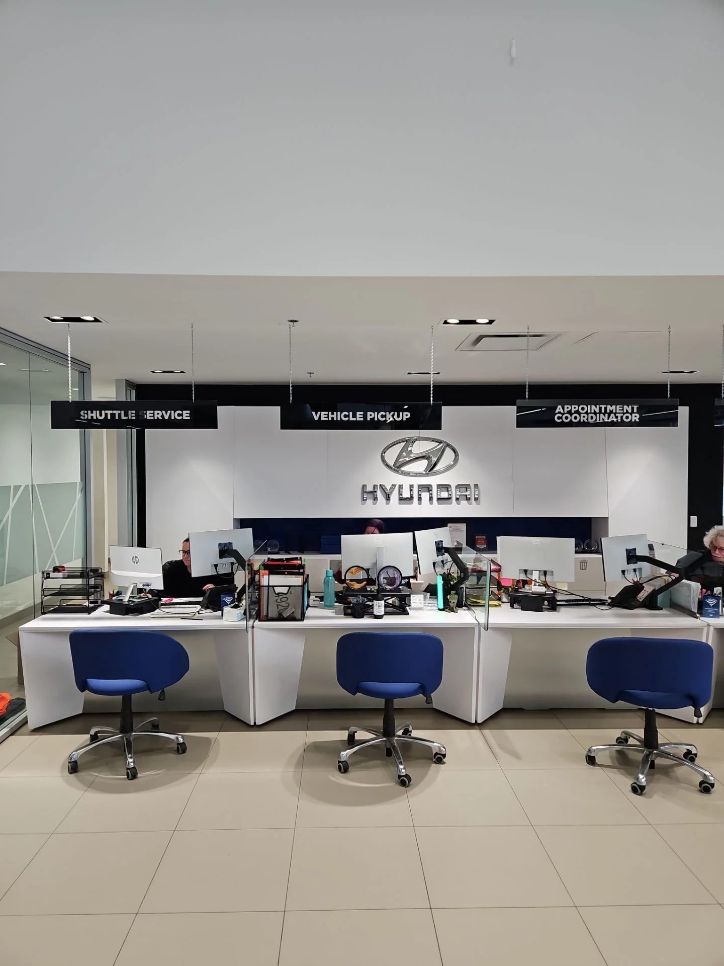 Hyundai service counter with three desks, each operated by staff, and blue chairs in front. Signs hanging above indicate shuttle service, vehicle pickup, and appointment coordinator.