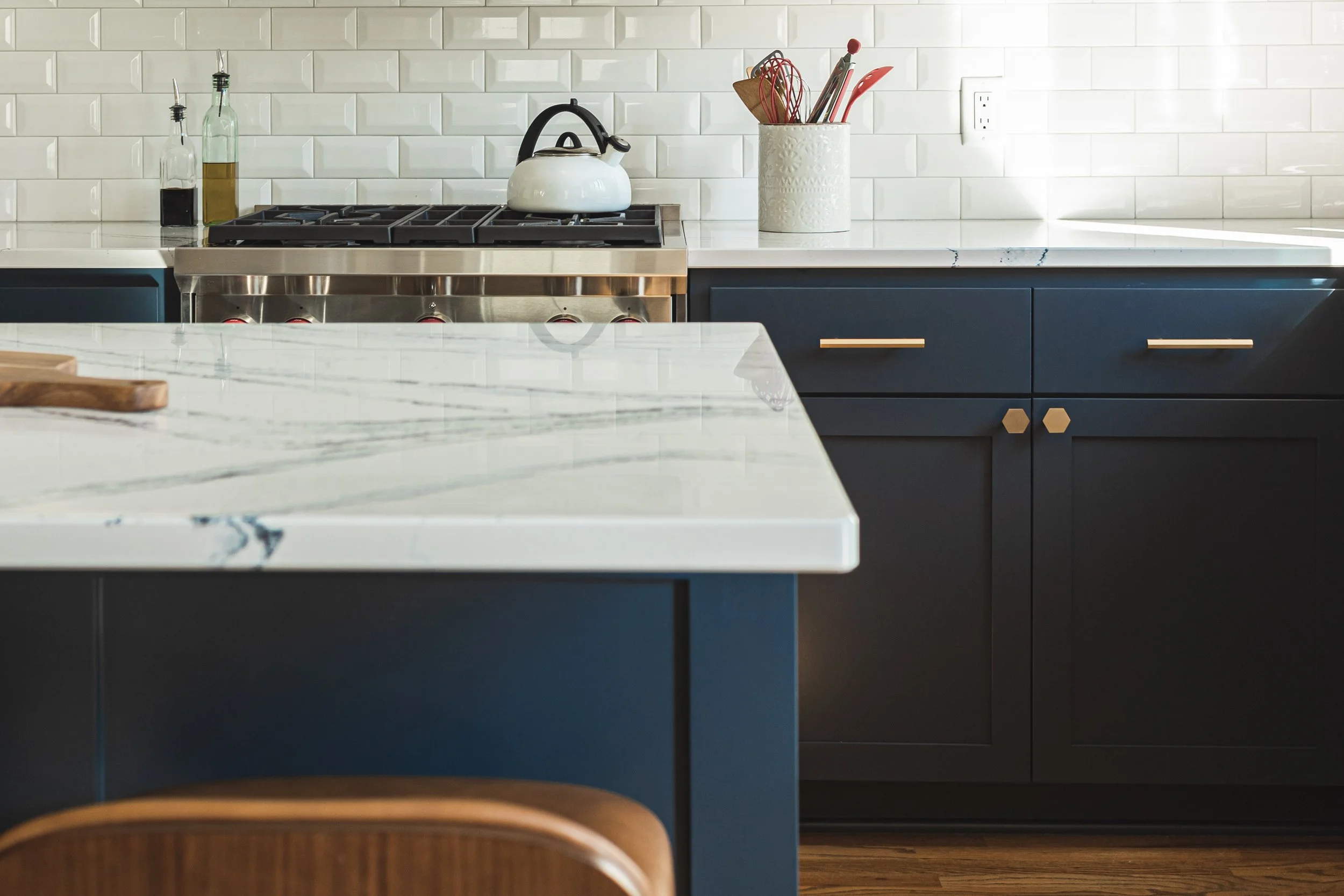 Modern kitchen featuring white tile backsplash, dark blue cabinets, marble countertops, stainless steel gas stove with a white kettle, and utensils in a holder.