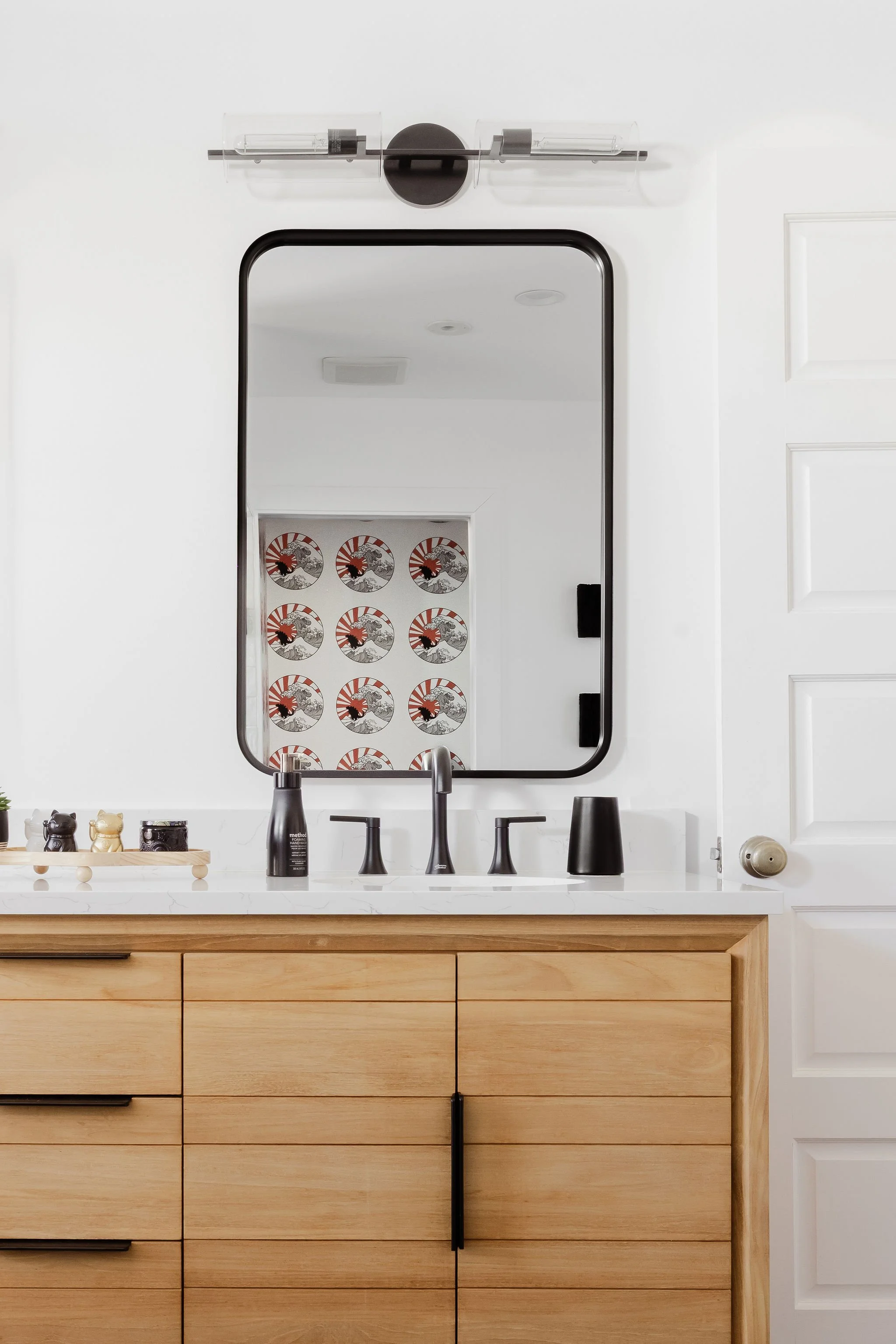 A bathroom vanity with a rectangular mirror above, black faucet, black soap dispenser, black toothbrush holder, and small decorative items on a wood cabinet, with a white wall and a light fixture above the mirror.