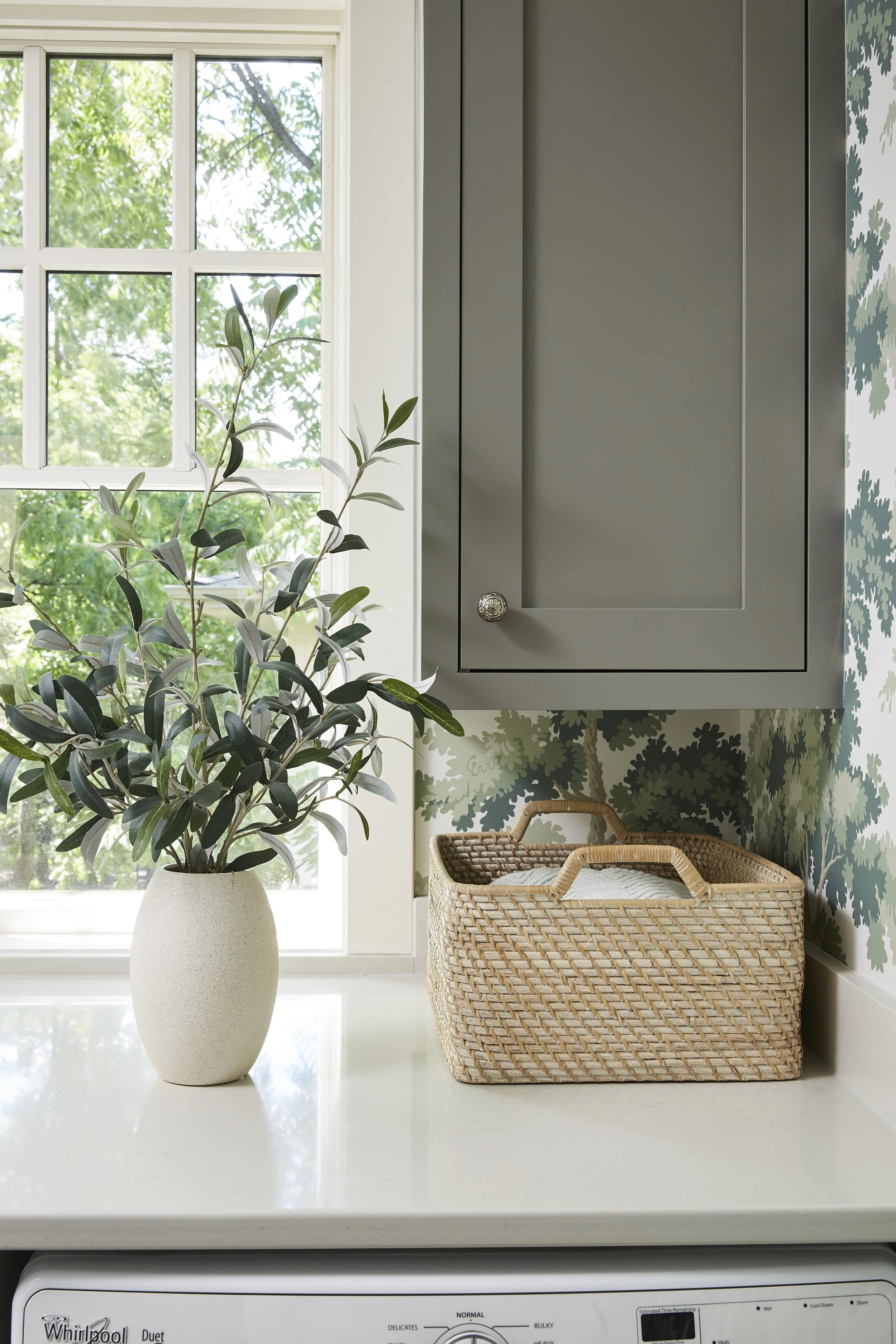 Laundry room with a white countertop, a vase with greenery, a wicker basket, a gray cabinet, and a window.
