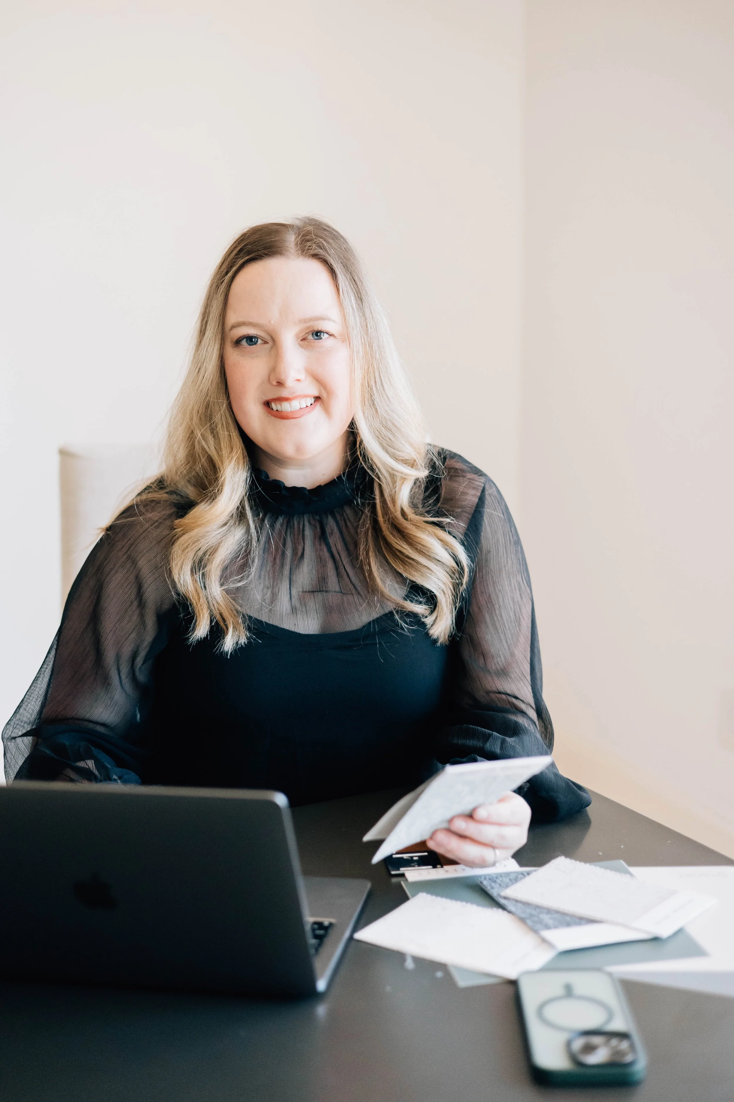 Woman with long blonde hair in a black blouse sitting at a desk with a laptop and holding fabric or paper samples.