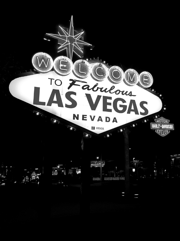 Night-time image of the iconic 'Welcome to Fabulous Las Vegas Nevada' sign with illuminated lights, including a star on top and a Harley-Davidson logo on the right.
