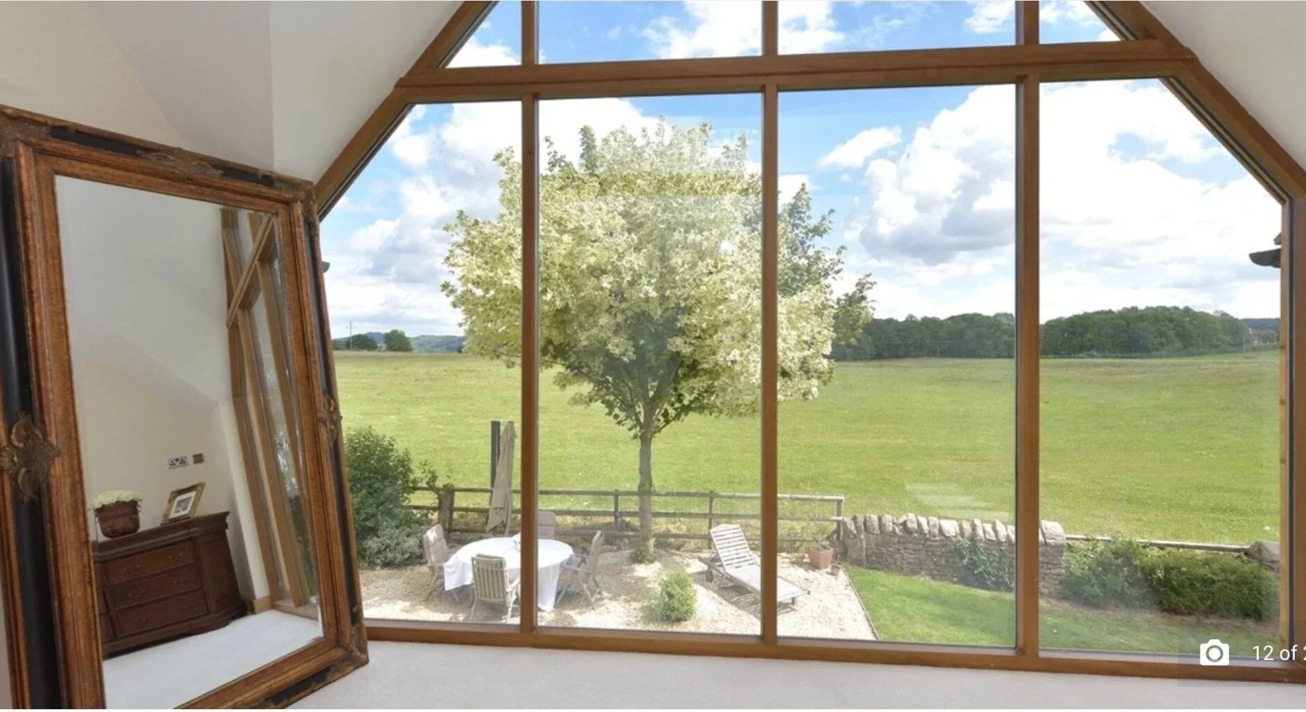 Interior of a room with a large window showing a green field, a tree, clouds, and a blue sky outside. A large, ornate gold-framed mirror leans against the wall, reflecting part of the room and the window outside.