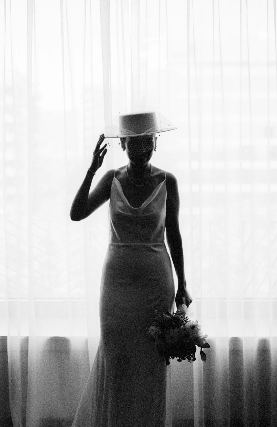 foto analógica en blanco y negro de una novia con sombrero delante de una ventana a contraluz