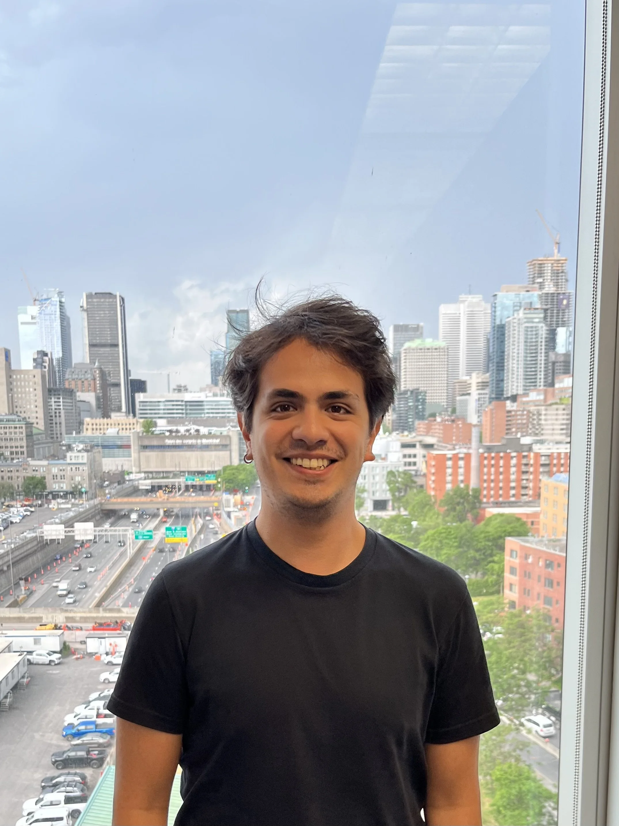 Smiling young man with dark hair wearing a black t-shirt standing in front of a large window overlooking a cityscape with tall buildings, roads, and trees.