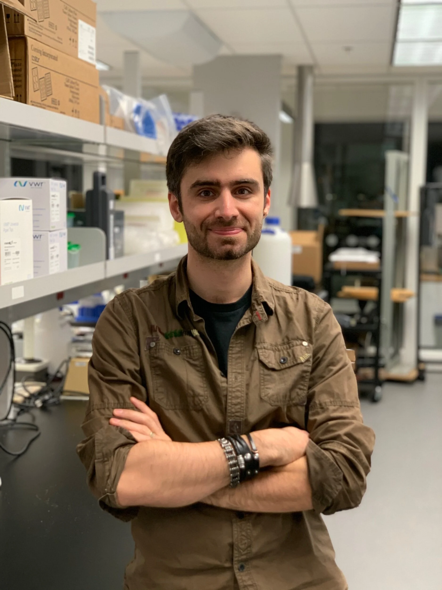 Young man with brown hair, beard, and bracelets on his wrist, standing in a laboratory with shelves of supplies in the background.