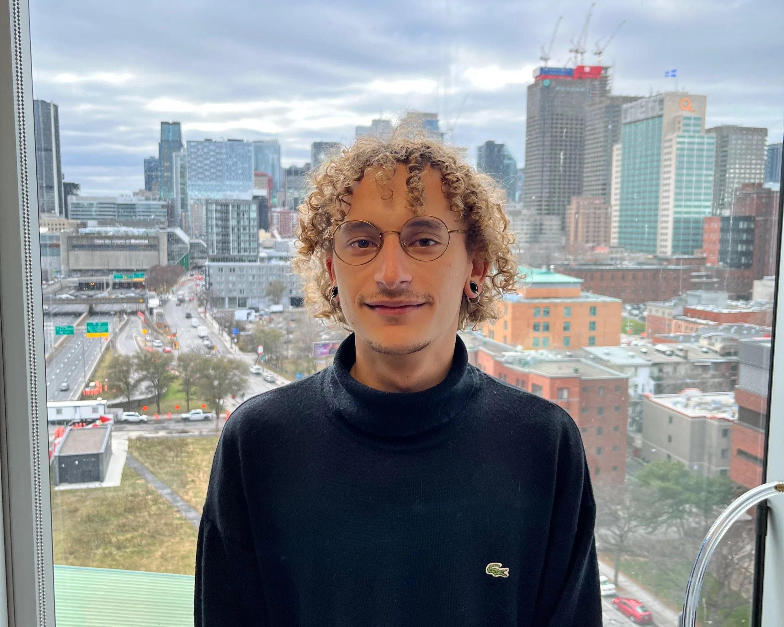 A young man with curly blonde hair, glasses, and earrings standing in front of a large window with a cityscape background of tall buildings and overcast sky.