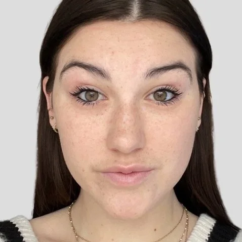 Close-up of a young woman with light skin, long dark hair, and light-colored eyes, looking directly at the camera, wearing a white and black top and small earrings.