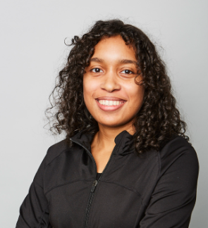 Portrait of a young woman with curly dark hair smiling, wearing a black jacket against a plain light gray background.