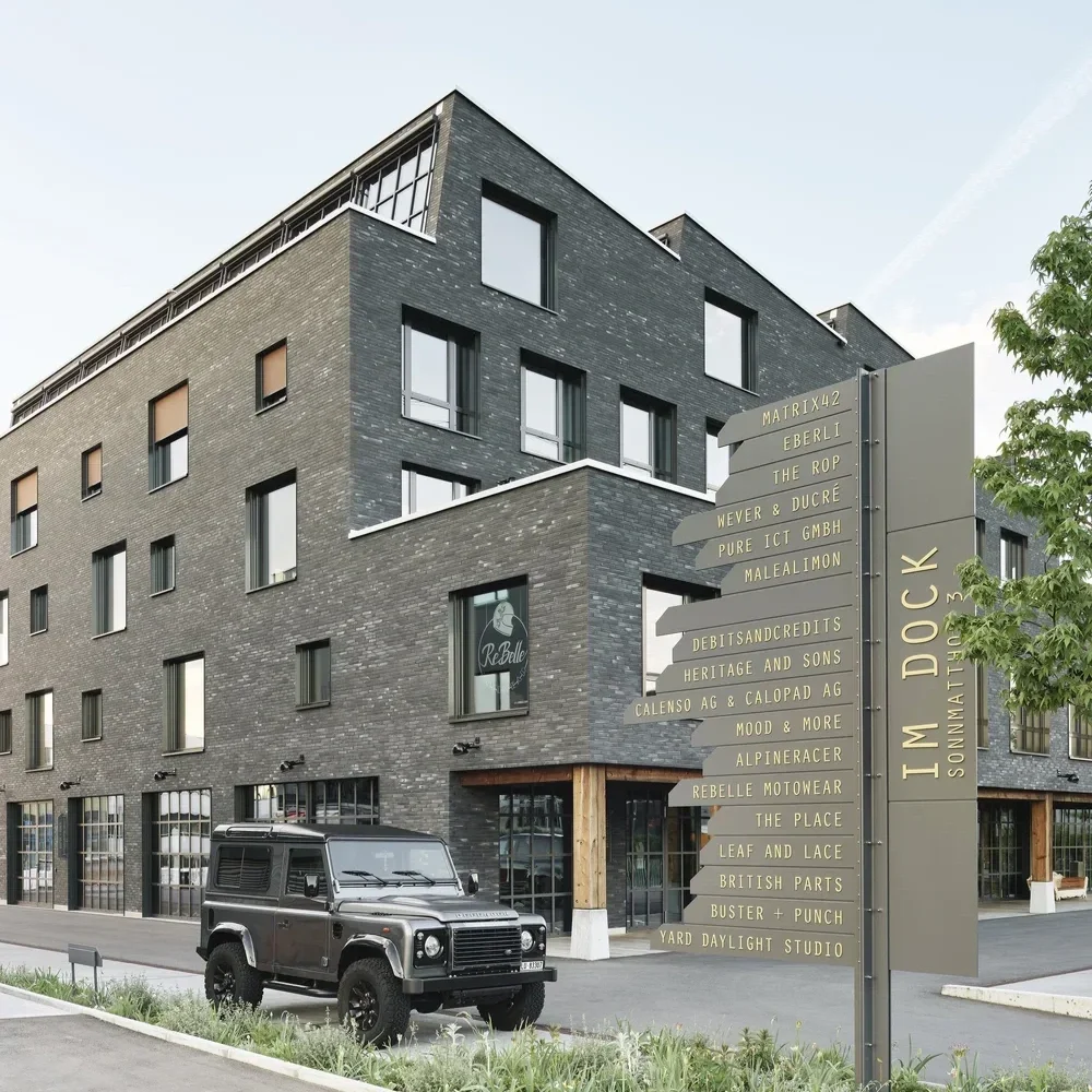Mehrstöckiges, modernes Gebäude mit dunklen Ziegelwänden und großen Fenstern. Es steht vor einer Straße mit einem schwarzen Geländewagen davor. Neben dem Gebäude befindet sich ein Wegweiser mit mehreren Richtungen und Namen, der auf den I' M DOC Sonntagsmarkt hinweist.