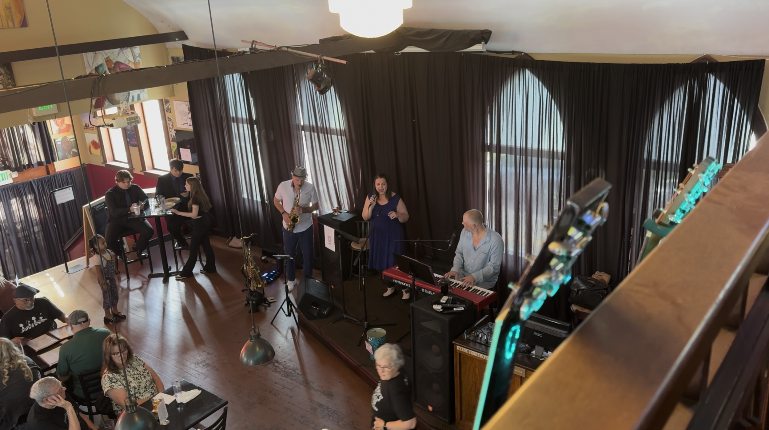 Jazz band performing on a stage with a woman singing, a man playing keyboard, and a saxophonist. Audience members seated at tables watch the performance in a venue with large windows, dark curtains, and hanging lights.