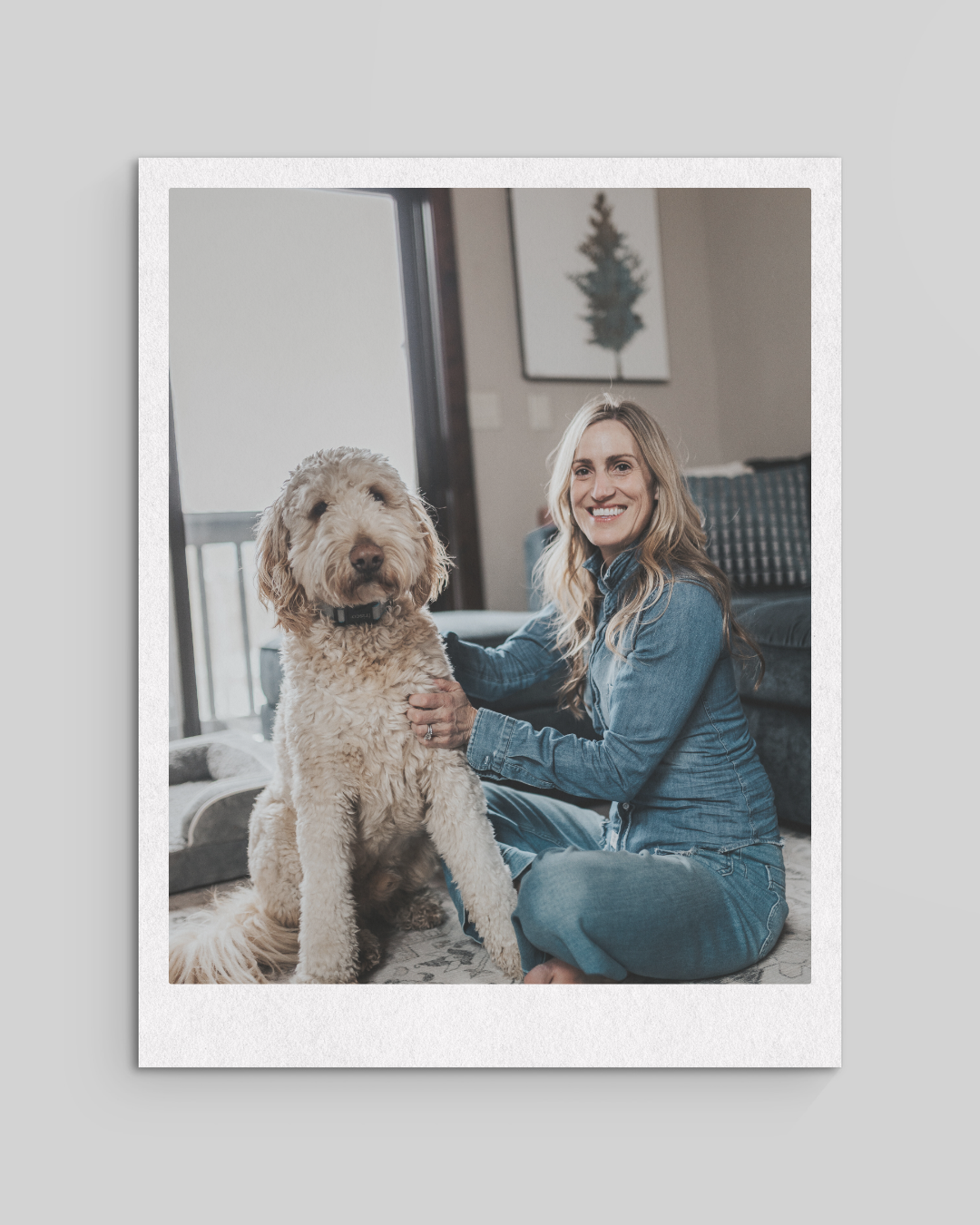 A woman sitting on a rug with a large, fluffy, cream-colored dog inside a living room, smiling at the camera.