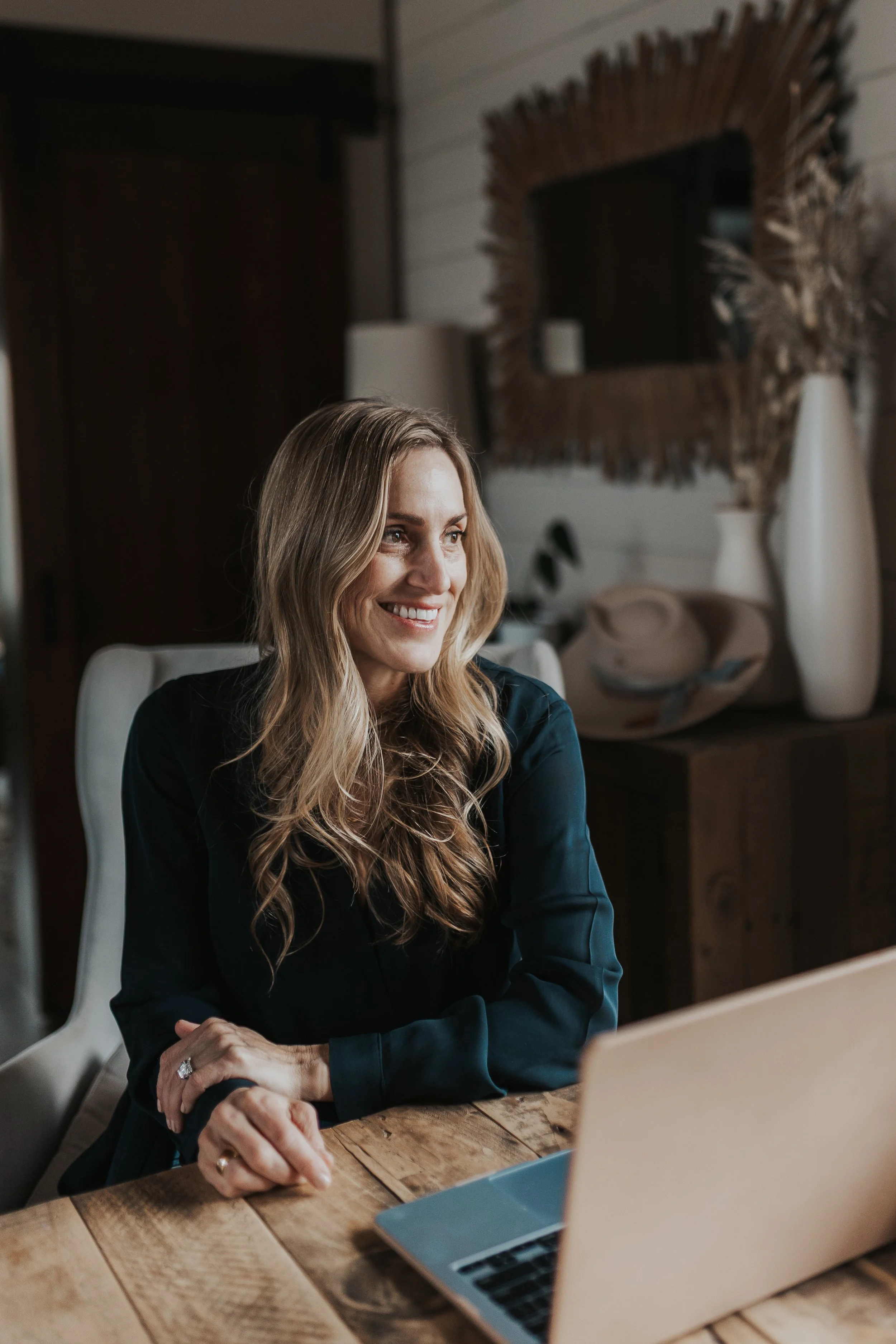 A woman with long, wavy blond hair smiling while sitting at a wooden table with a laptop in front of her.