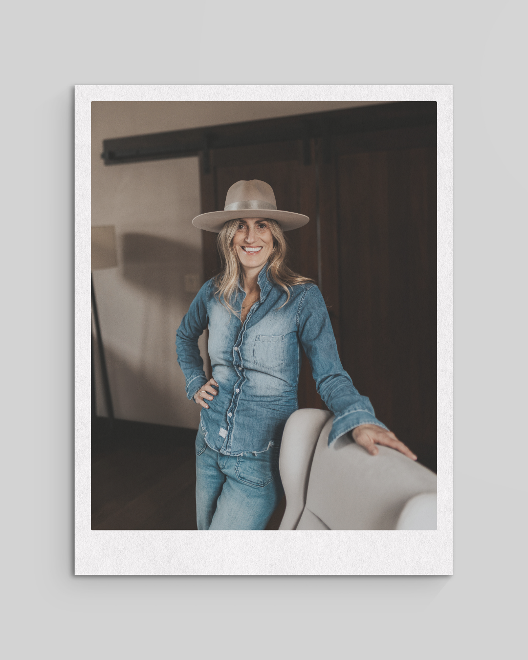 Woman wearing a wide-brimmed hat and denim outfit smiling while leaning on a chair indoors.