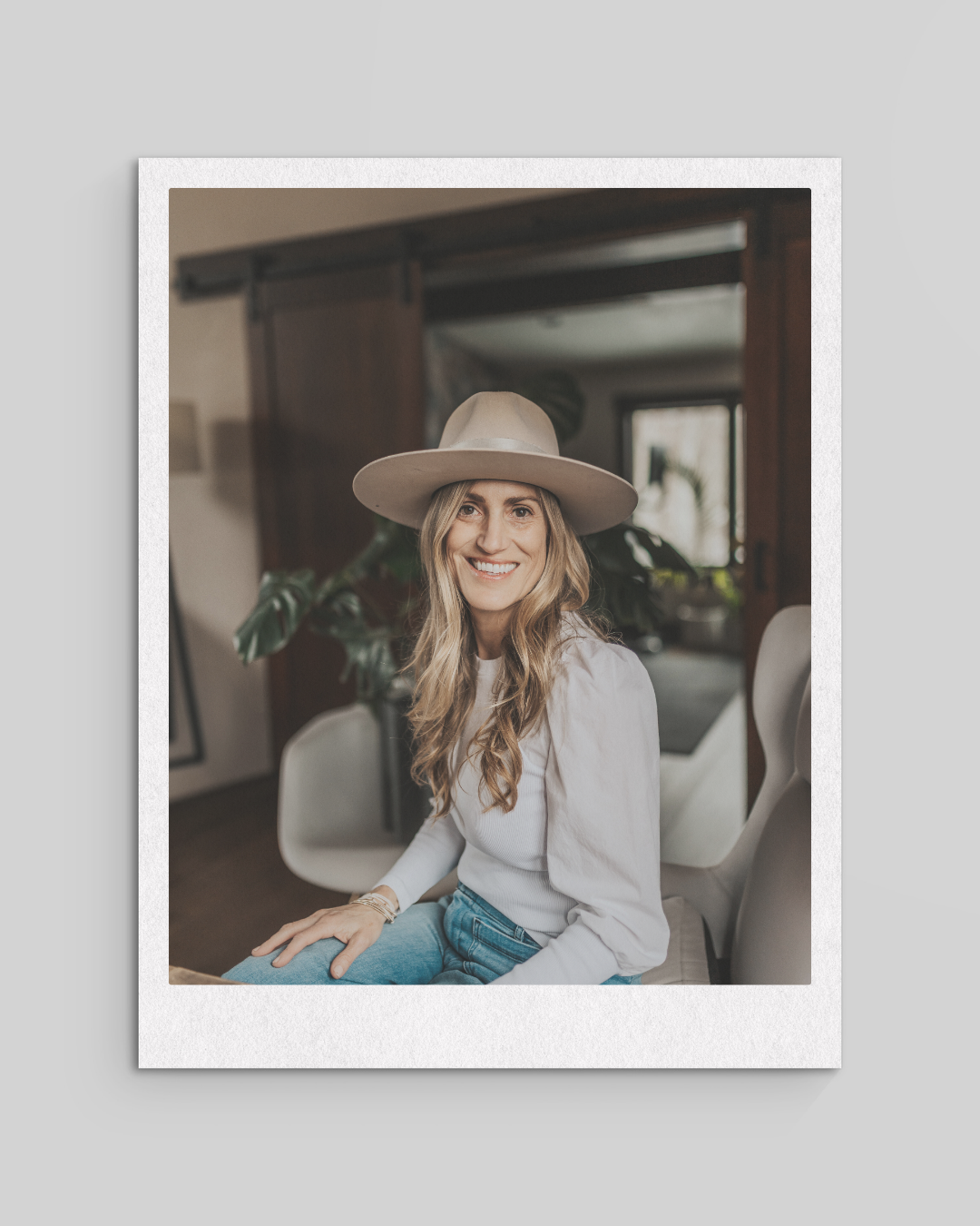 A woman with long blond hair, wearing a large beige hat and white top, sitting indoors on white chairs, smiling at the camera with a houseplant and window in the background.