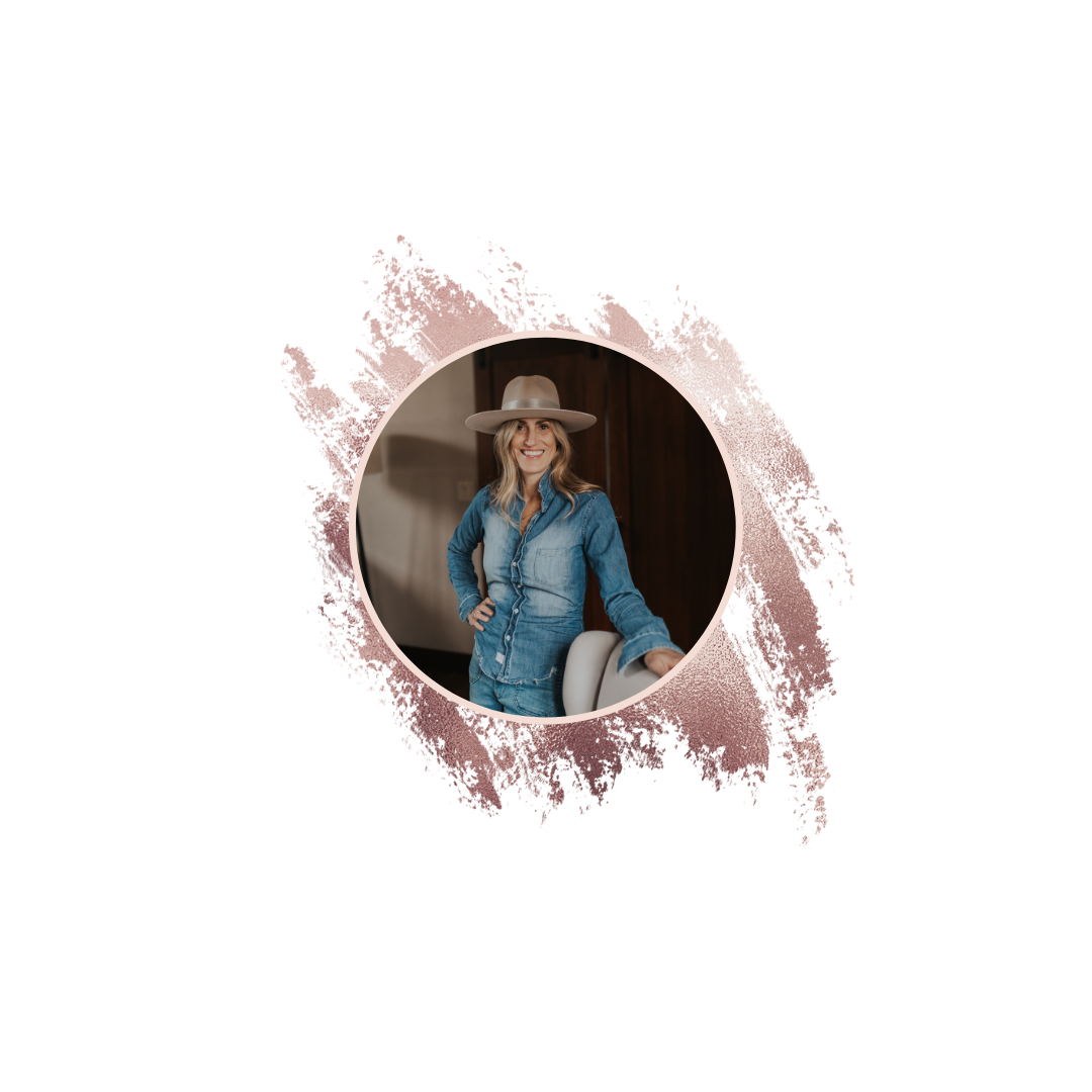 A woman wearing a wide-brimmed hat and denim outfit, smiling and standing indoors near a chair.