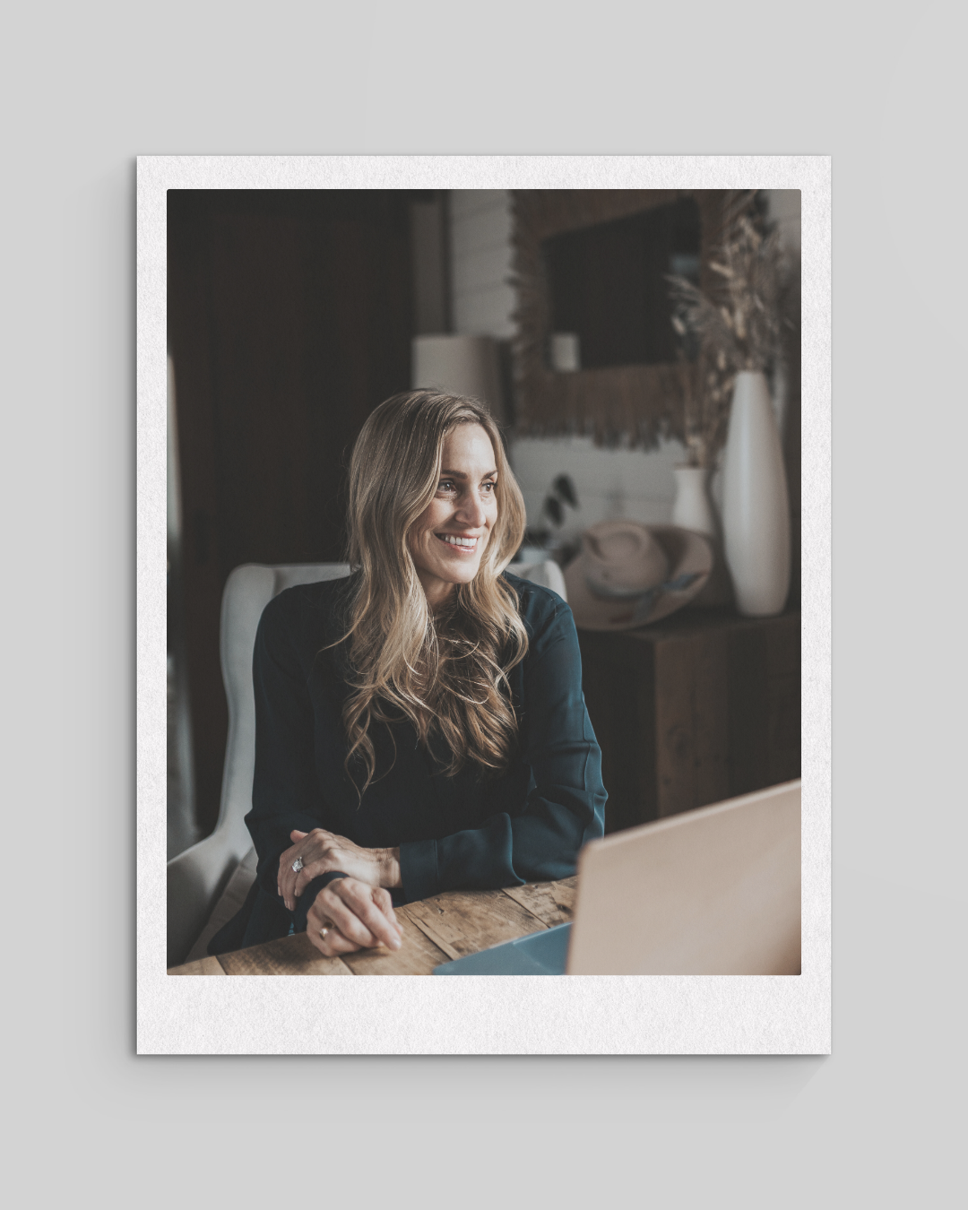A smiling woman with long wavy blonde hair sitting at a wooden table, using a laptop, in a cozy, stylish room with decorative vases and dried plants in the background.