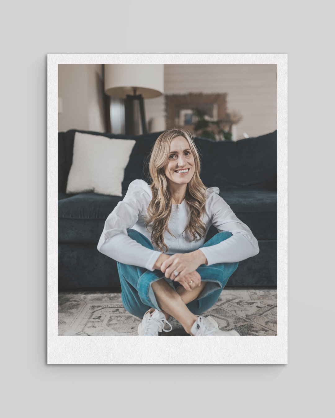 A woman sitting on the floor in a living room, smiling, wearing a white shirt and blue jeans.