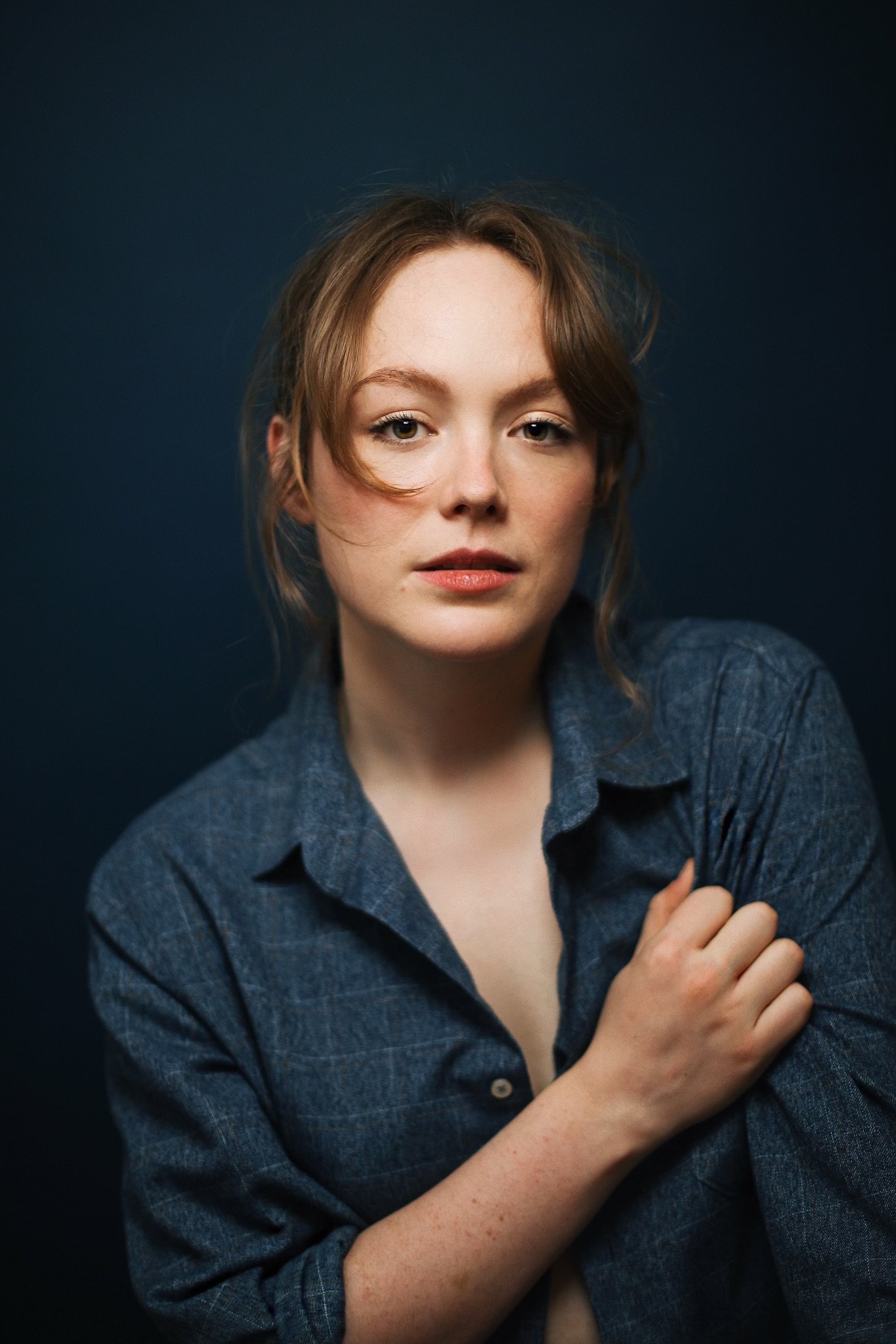 Holly Hinchliffe with light brown hair and fair skin wearing a dark blue plaid shirt, looking at the camera with a neutral expression, posing against a dark background.